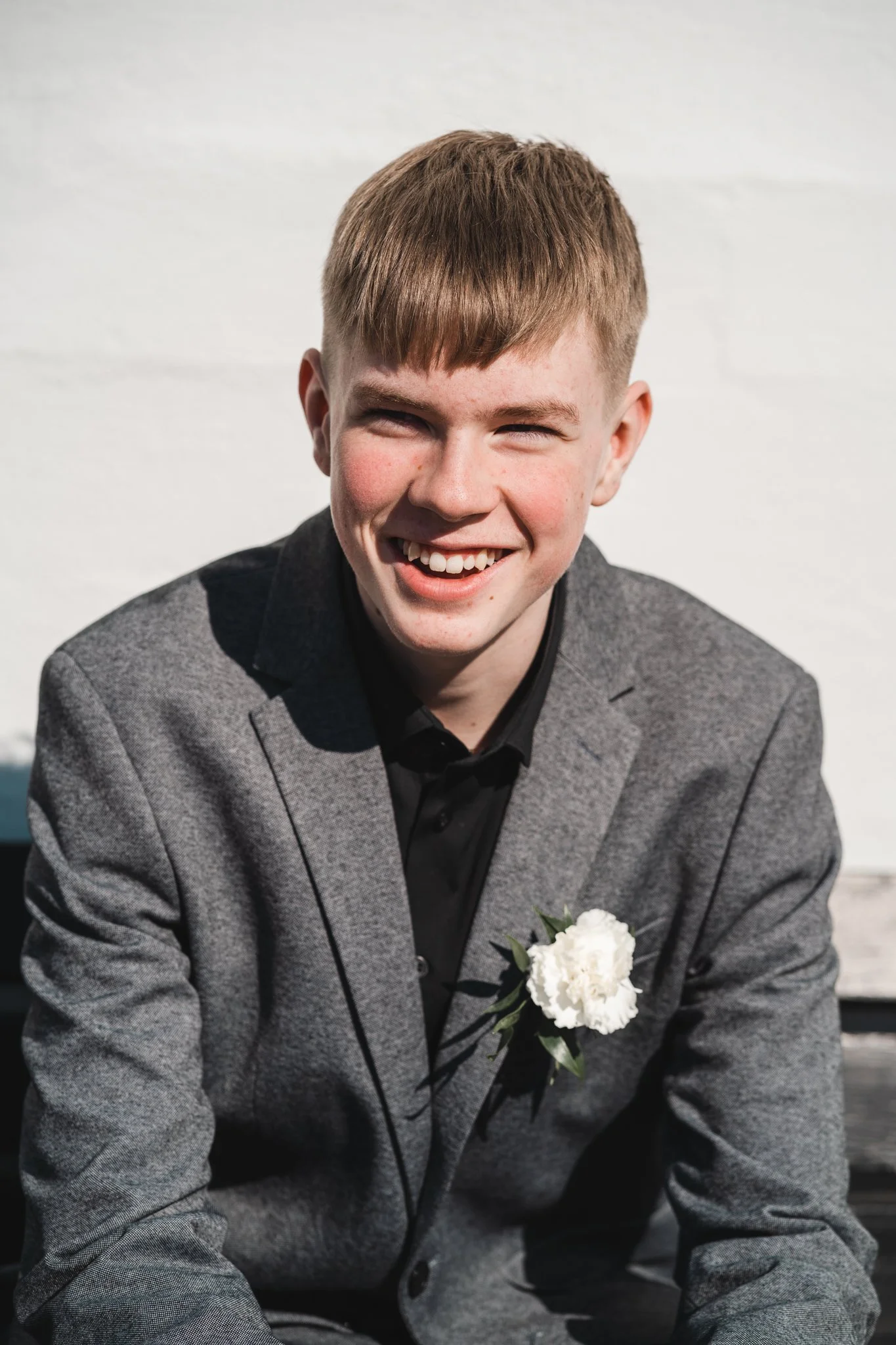 A young man with short brown hair, missing a front tooth, wearing a gray blazer with a white flower pinned to it, smiling outdoors against a white wall.