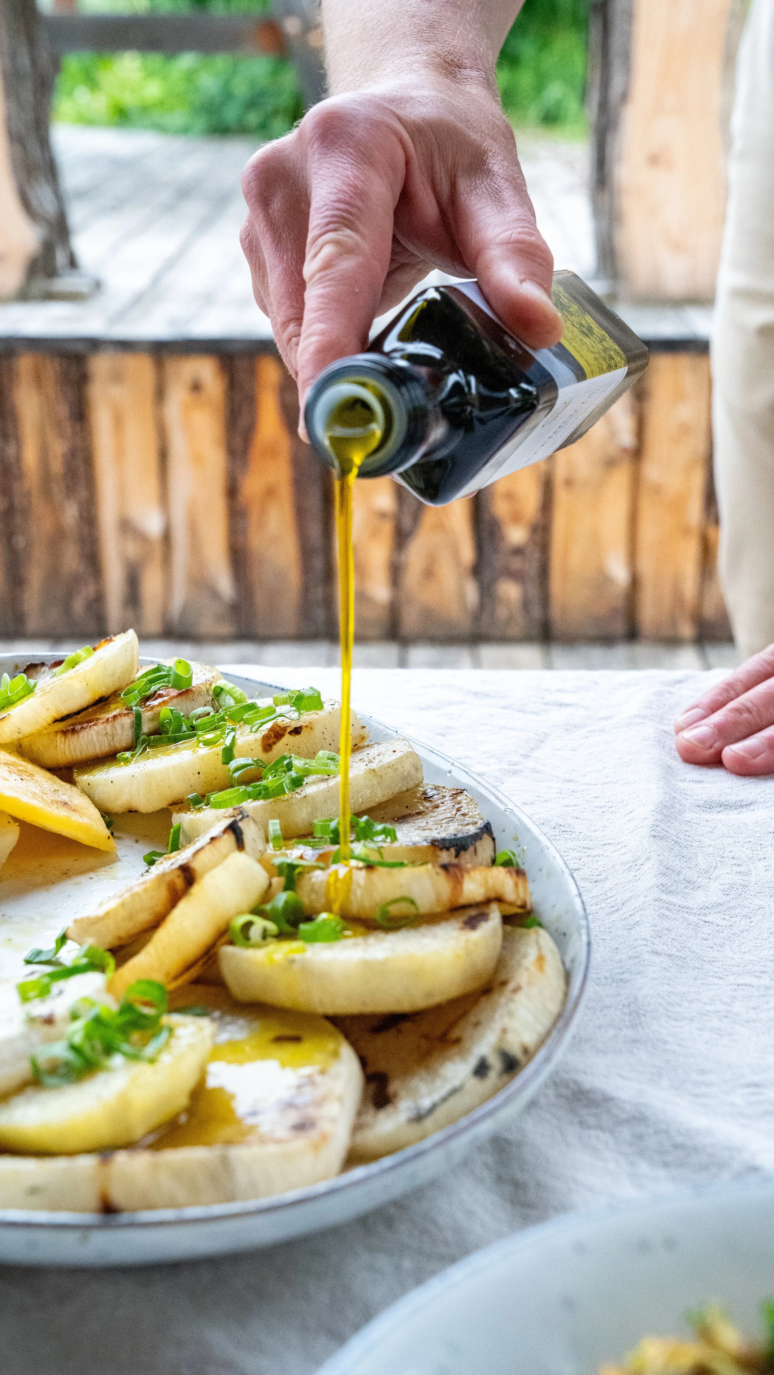 A person drizzling olive oil over a platter of grilled vegetables and lemon slices on a table with a white cloth, outdoors with a wooden background and green foliage.