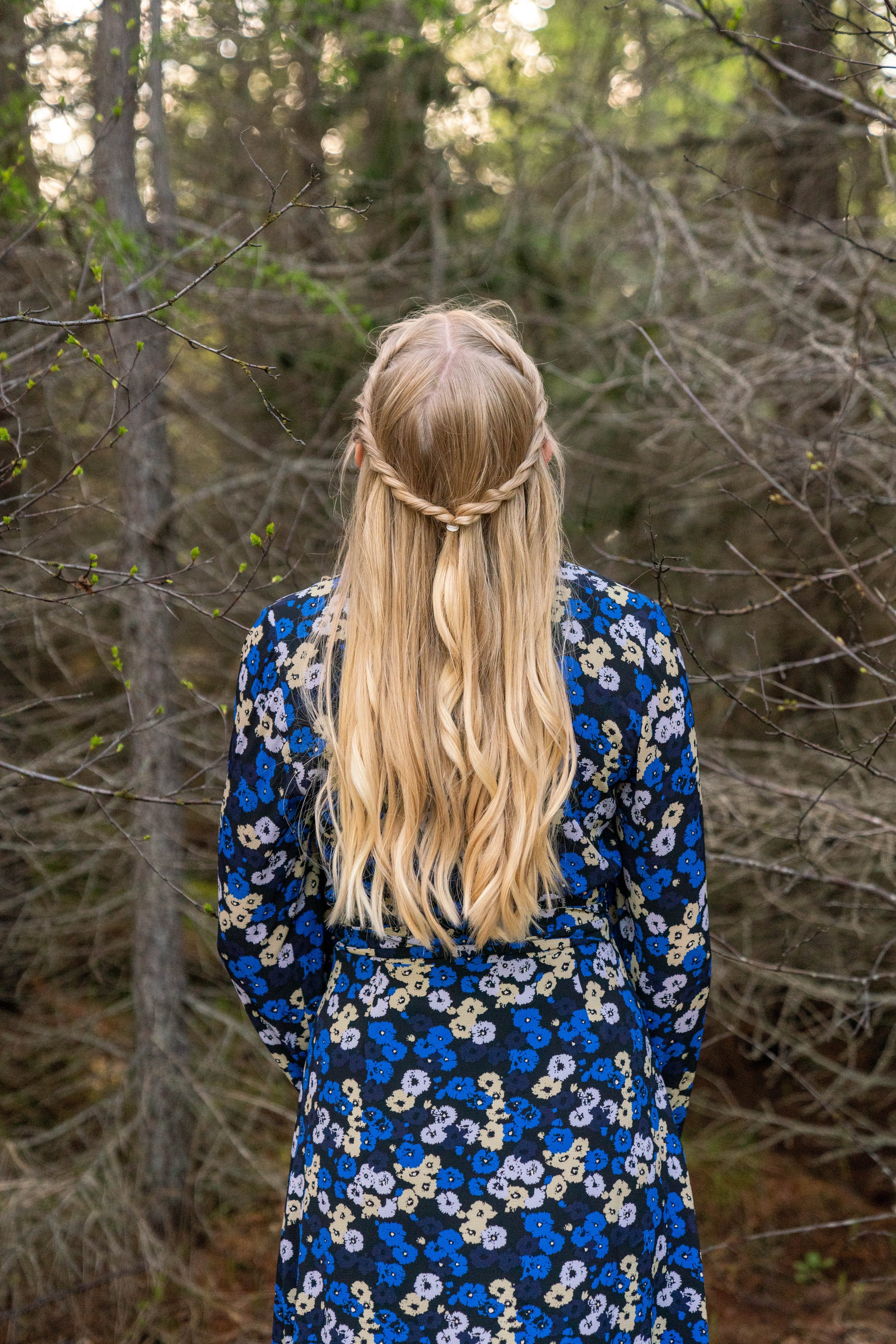 A woman with long blonde hair styled with a small braid on each side, wearing a blue floral dress, standing in a wooded area with bare and budding trees.