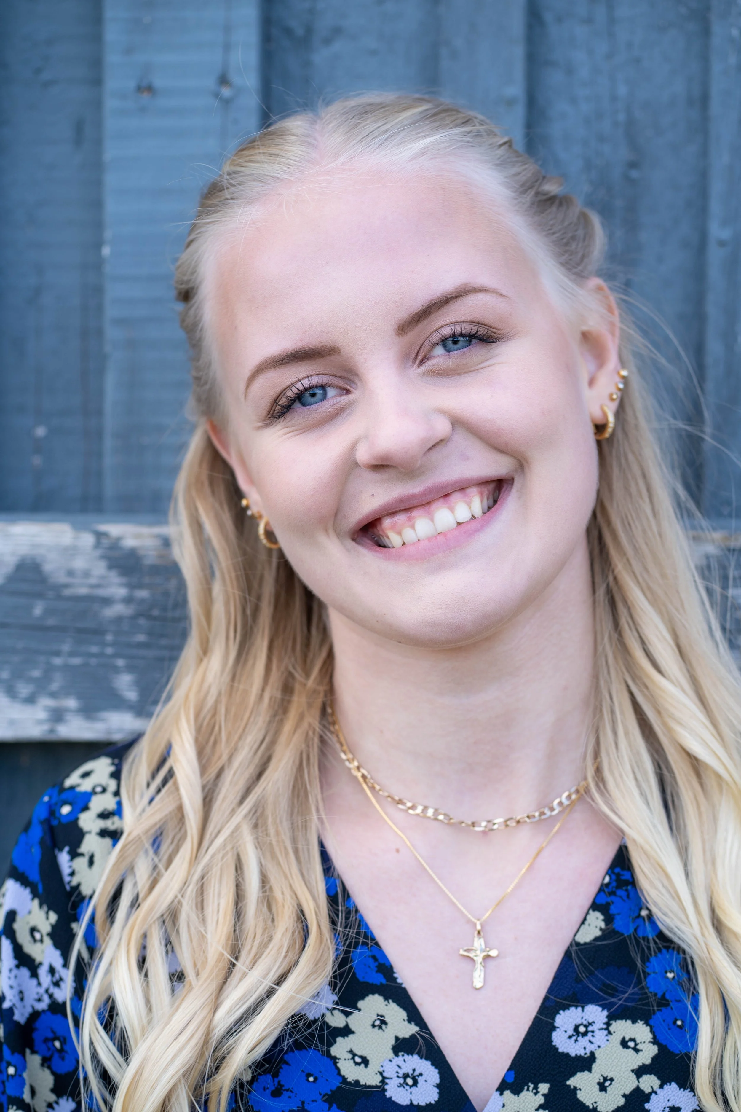 A young woman with long blonde hair, blue eyes, and a bright smile, wearing gold jewelry including hoop earrings, layered necklaces with a cross, and a floral-patterned top in front of a weathered blue wooden background.