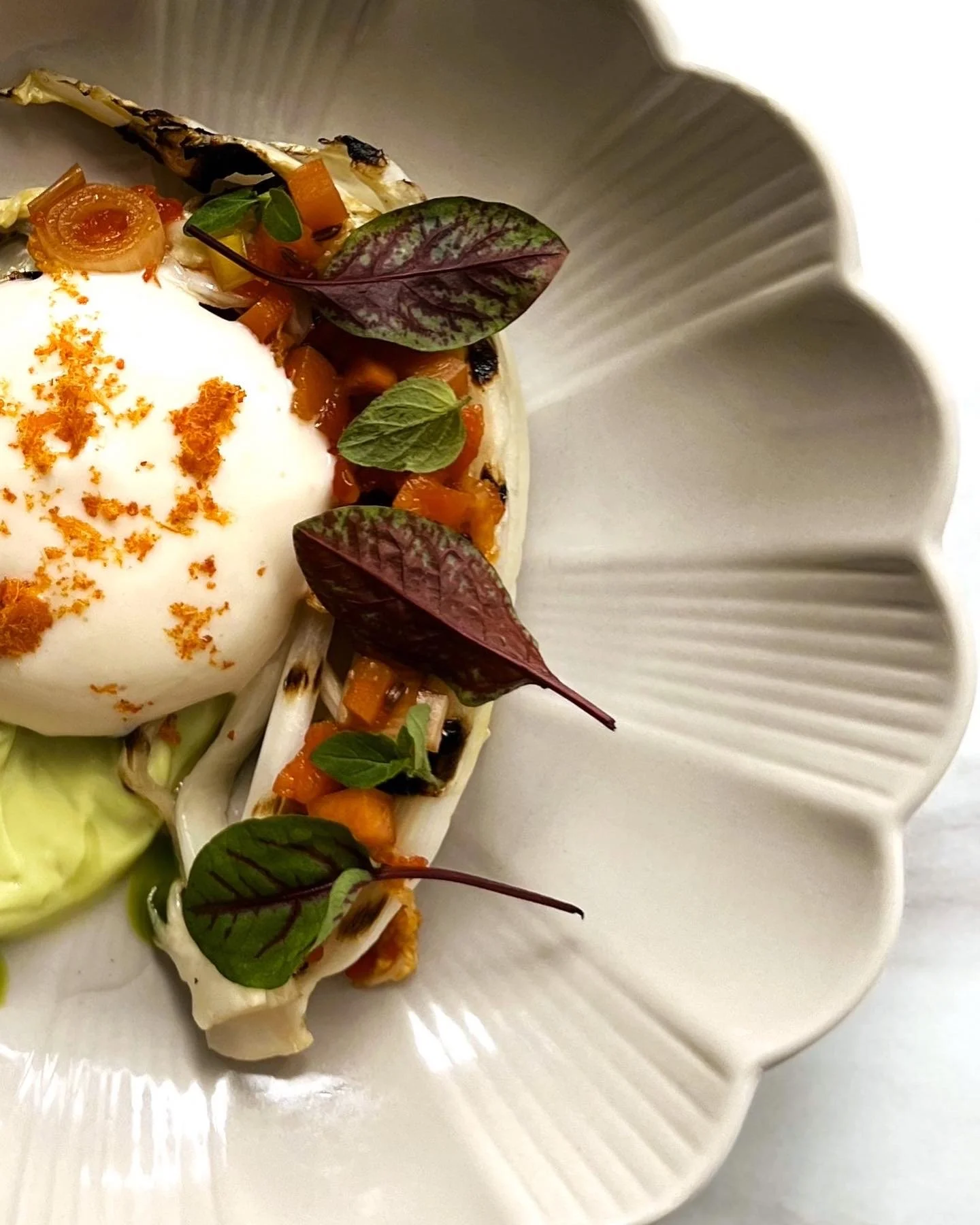 Poached egg topped with orange zest, served on a bed of grilled vegetables, with fresh herbs and edible leaves, on a decorative white plate.
