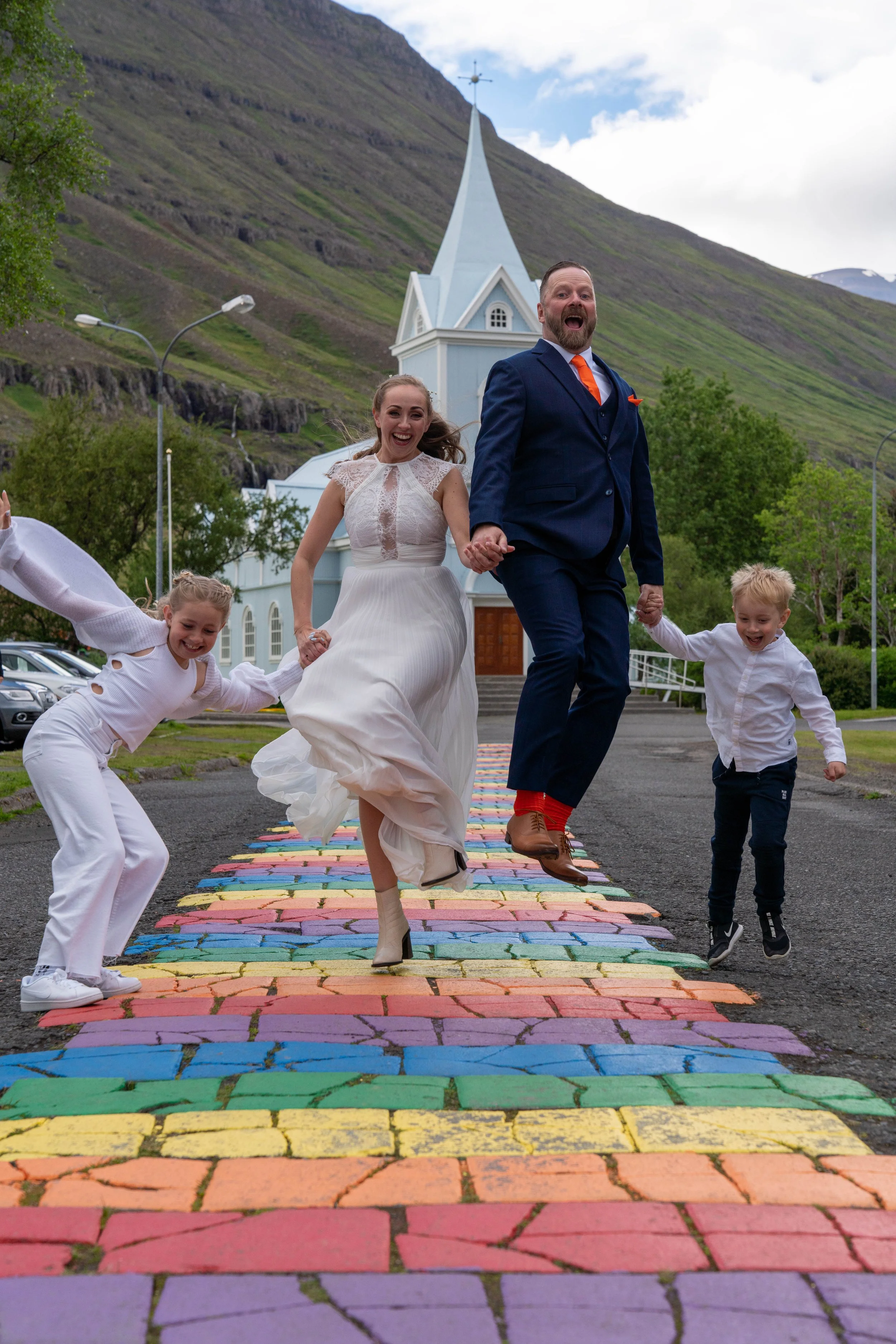 A couple dressed as bride and groom happily jumping on a colorful rainbow-painted crosswalk, holding hands with two children, in front of a small white church with a tall steeple, set against a green hillside with cloudy sky.