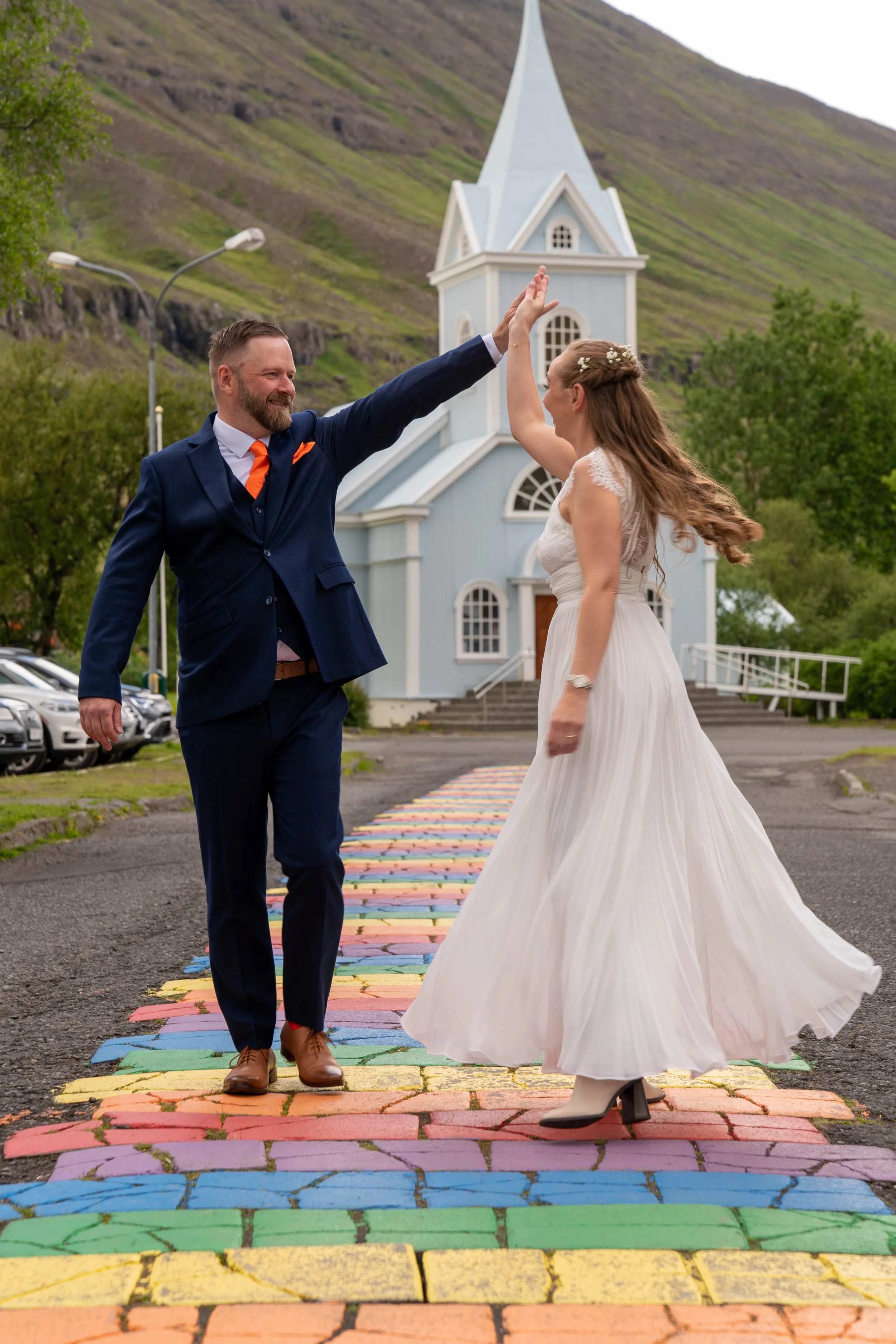 A bride and groom dancing on a colorful chalk-painted street in front of a church, with a green hillside in the background.