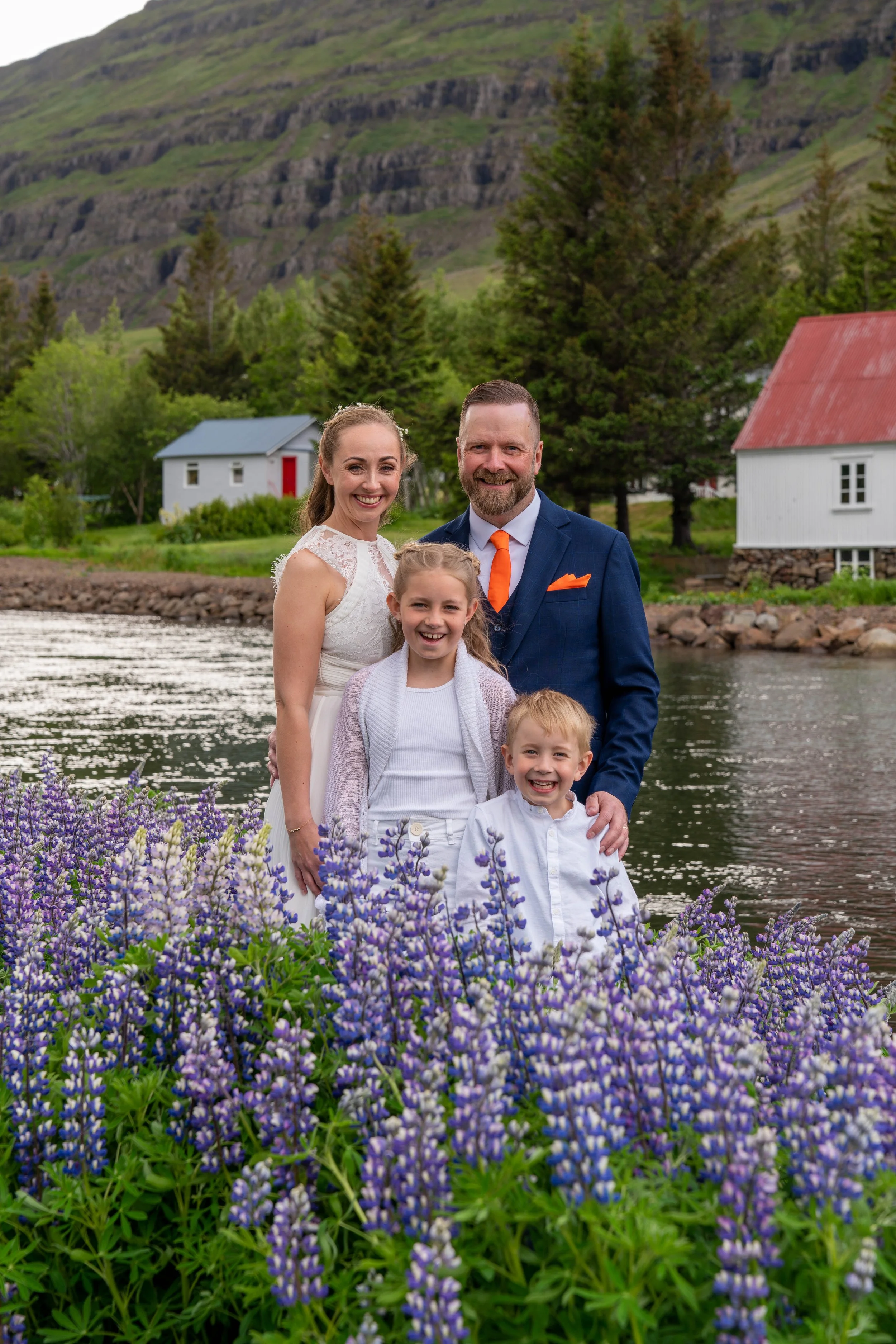 Family of four standing outdoors by a river with purple flowers in the foreground, a green hillside, trees, and white houses with red and blue roofs in the background, during daytime.