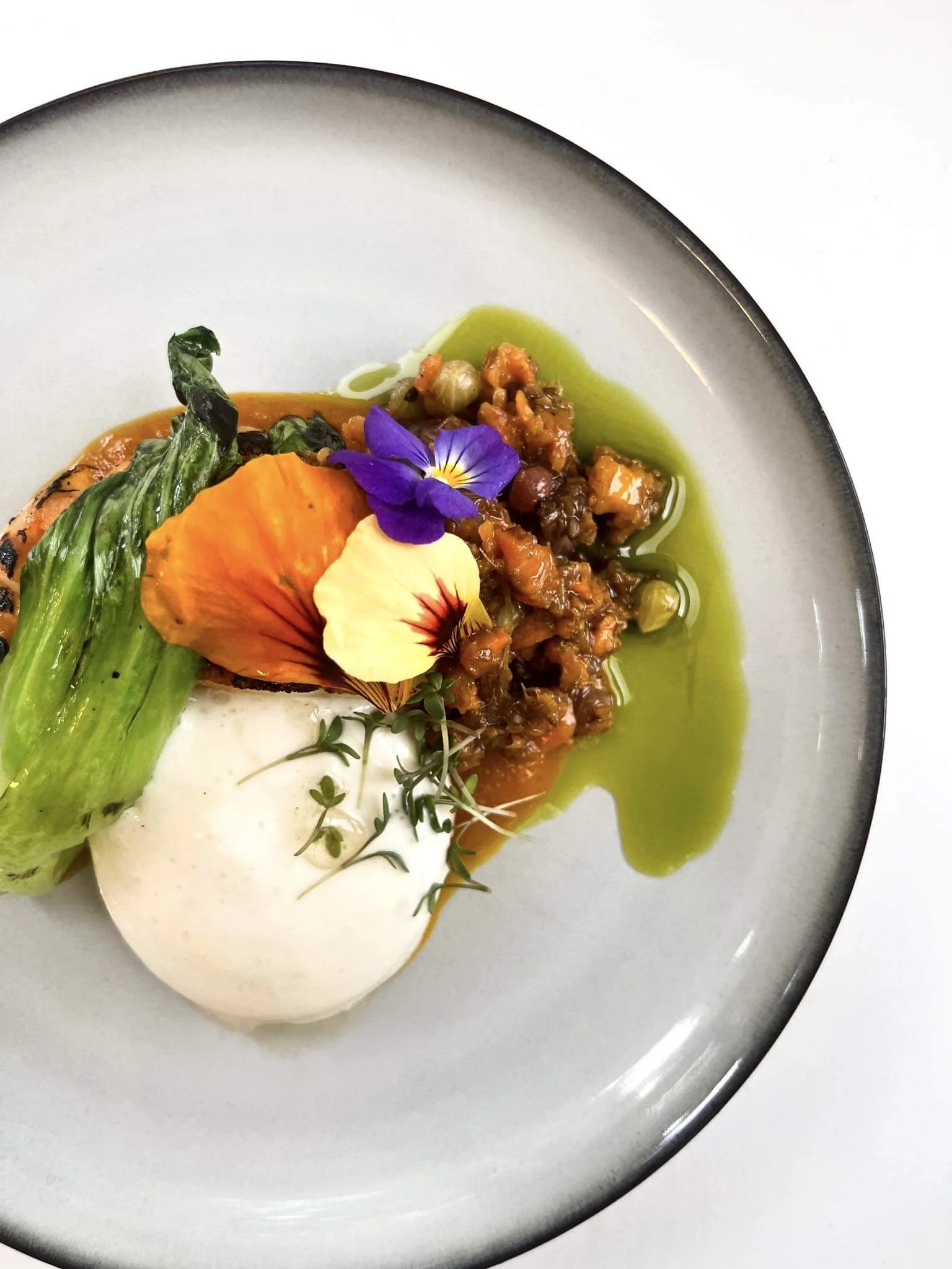 Gourmet plated dish with grilled green vegetable, carrot, lentil mixture, dollop of white sauce, microgreens, and edible flowers, garnished with green oil on a white plate with a black rim.