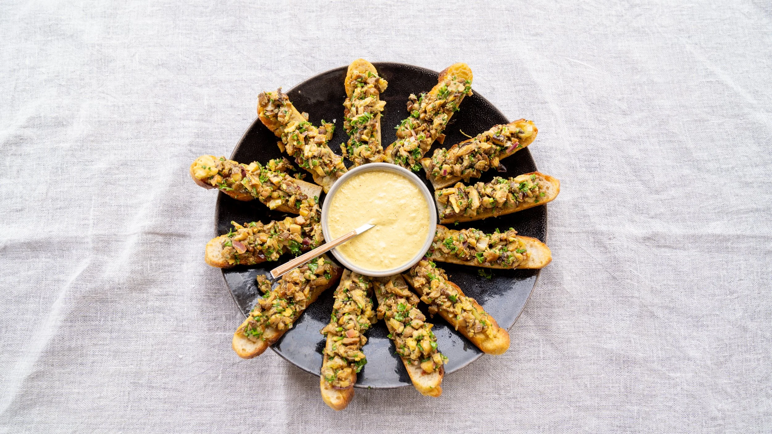 A black plate with toasted baguette slices topped with chopped vegetables and herbs, arranged around a small bowl of yellow dipping sauce, on a light gray tablecloth.