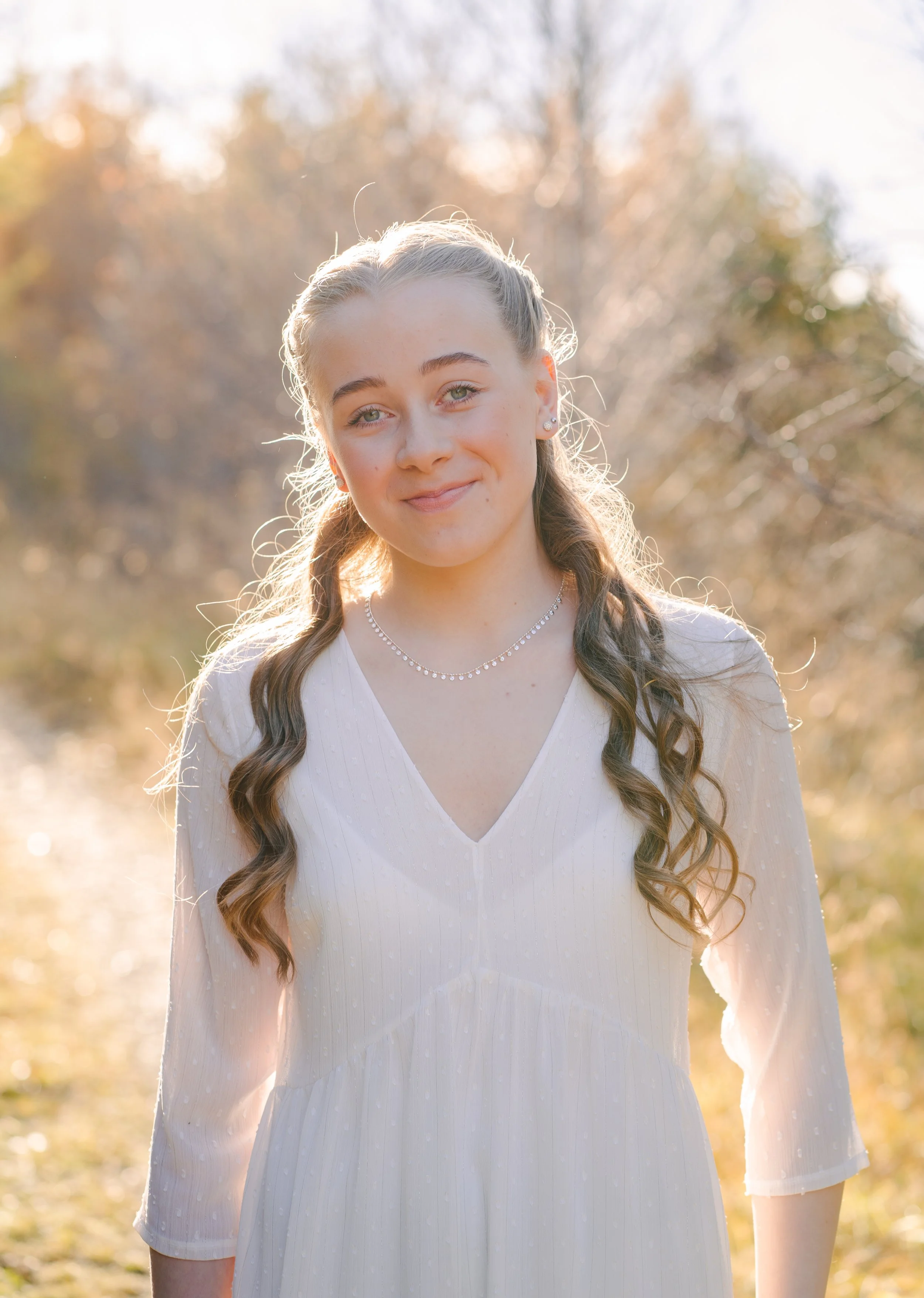 A young woman with long, wavy brown hair in a braid, wearing a white dress with three-quarter sleeves, standing outdoors in sunlight with a blurred background of trees and leaves.