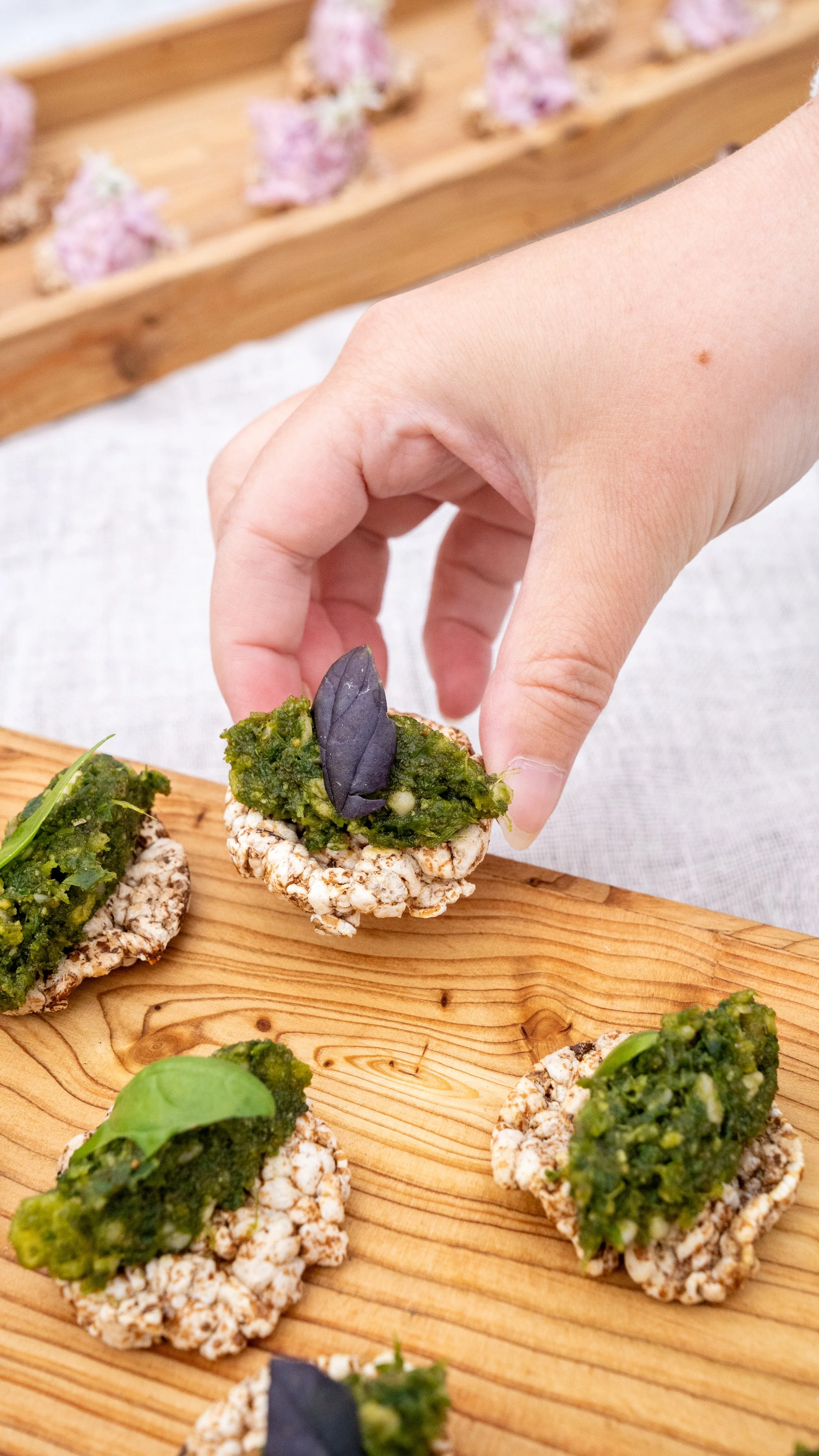 Close-up of a hand placing topped rice crackers with green spread and a purple basil leaf on a wooden serving board, with more rice crackers and a tray of pink and purple flowers in the background.