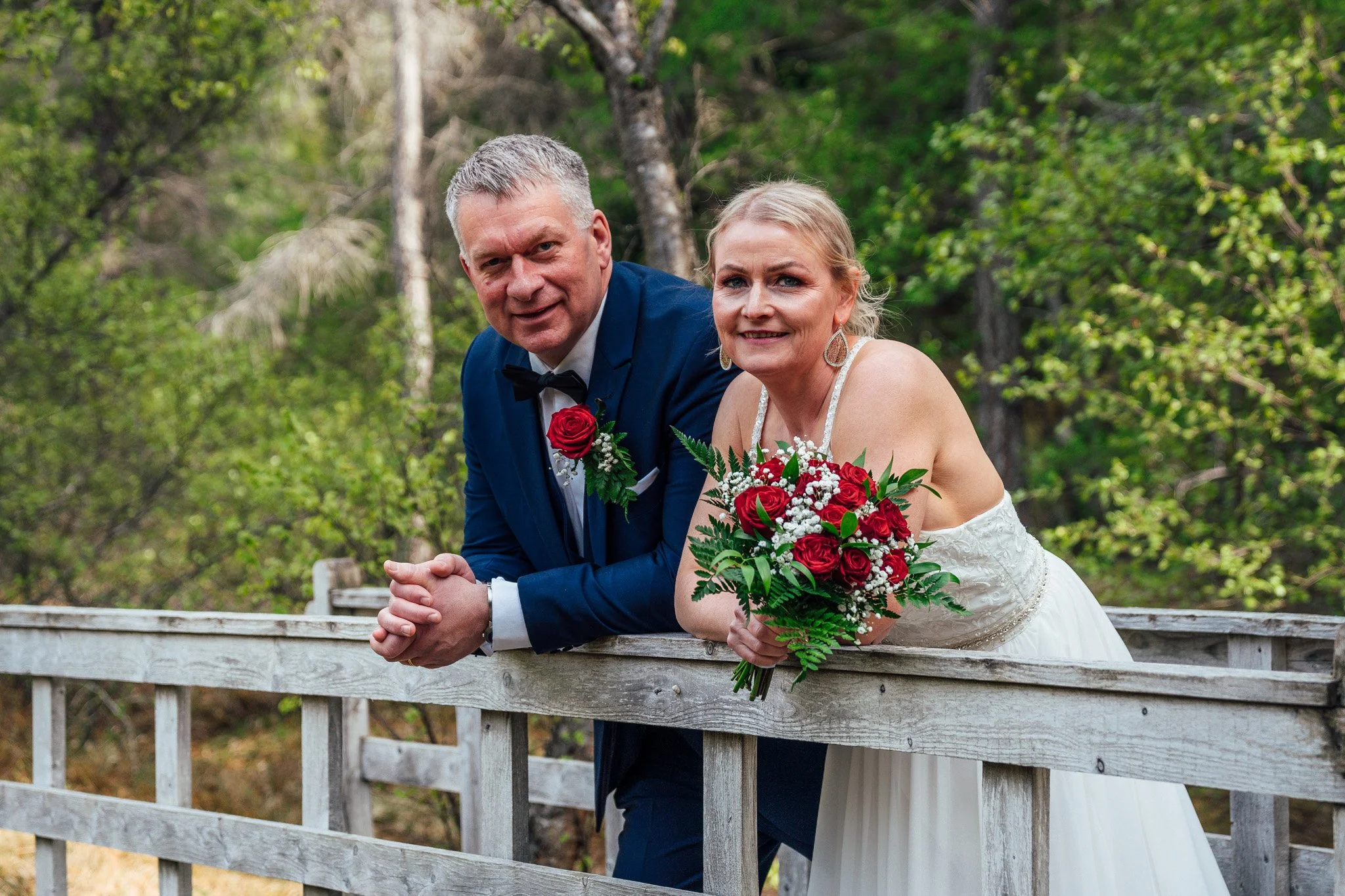 A bride and groom leaning on a wooden rail outdoors, surrounded by green trees, with the bride holding a bouquet of red and white flowers.