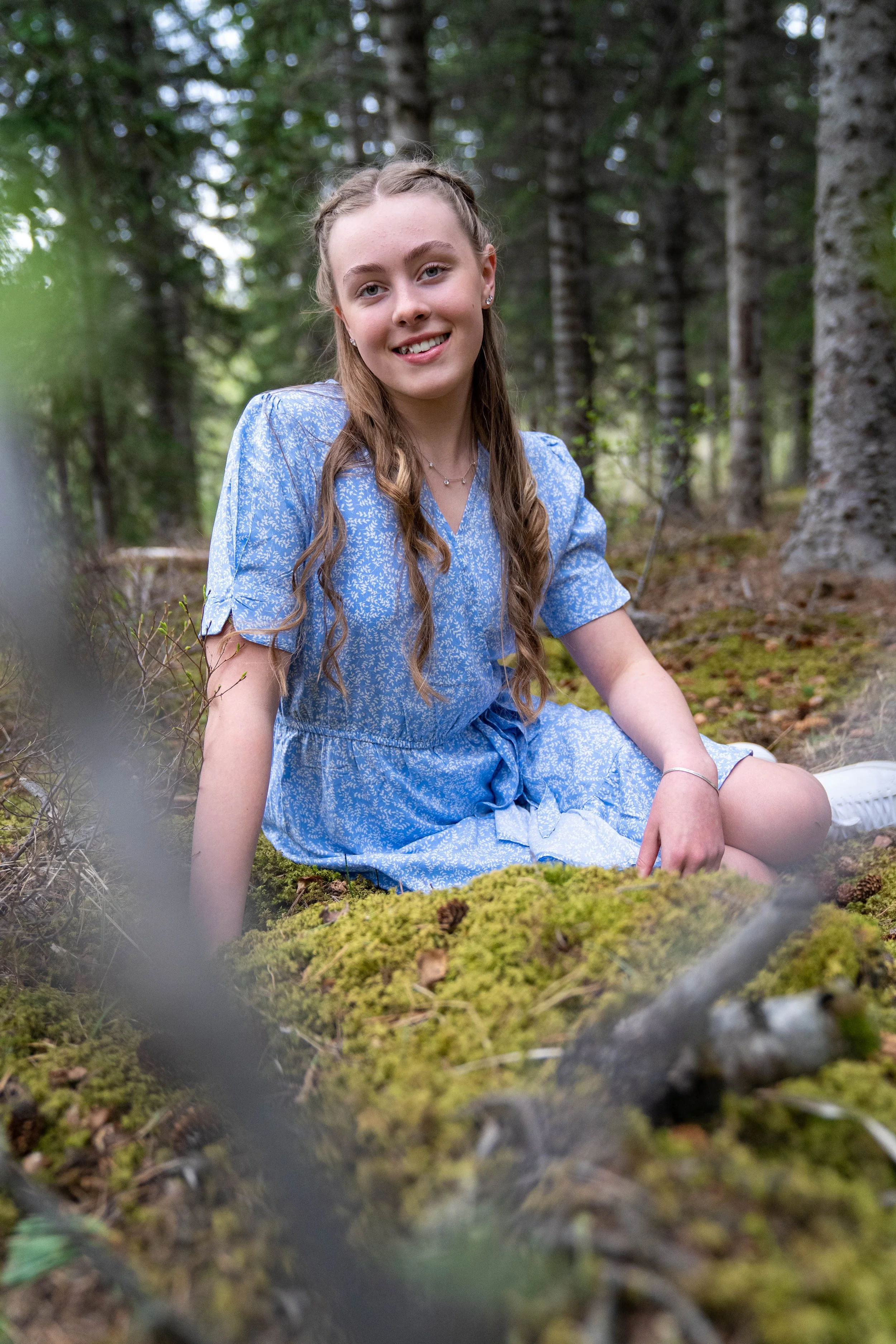 Young woman sitting on moss-covered ground in a forest, smiling at the camera, wearing a blue dress with a white pattern.