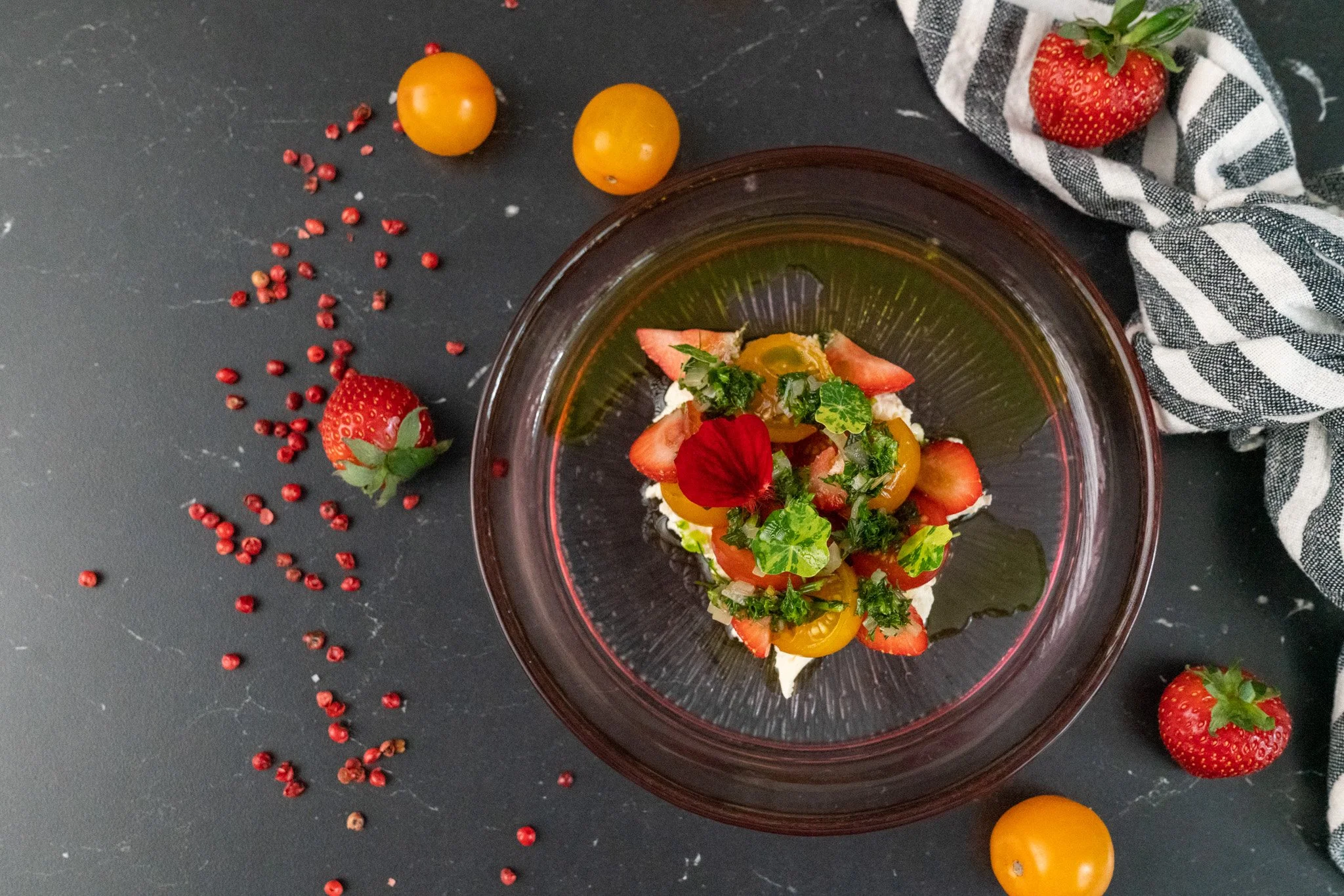 A dessert topped with strawberries, yellow and red cherry tomatoes, leafy greens, and an edible flower in a glass bowl. Whole strawberries, cherry tomatoes, and pink peppercorns are scattered on a dark surface, with a black-and-white striped cloth ne