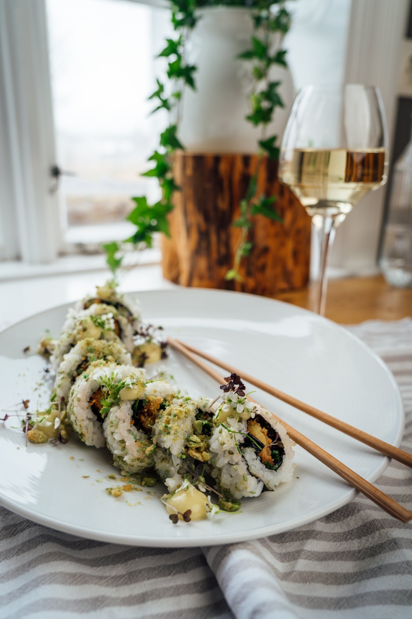 Sushi rolls with chopsticks, a glass of white wine, and a potted plant in the background.