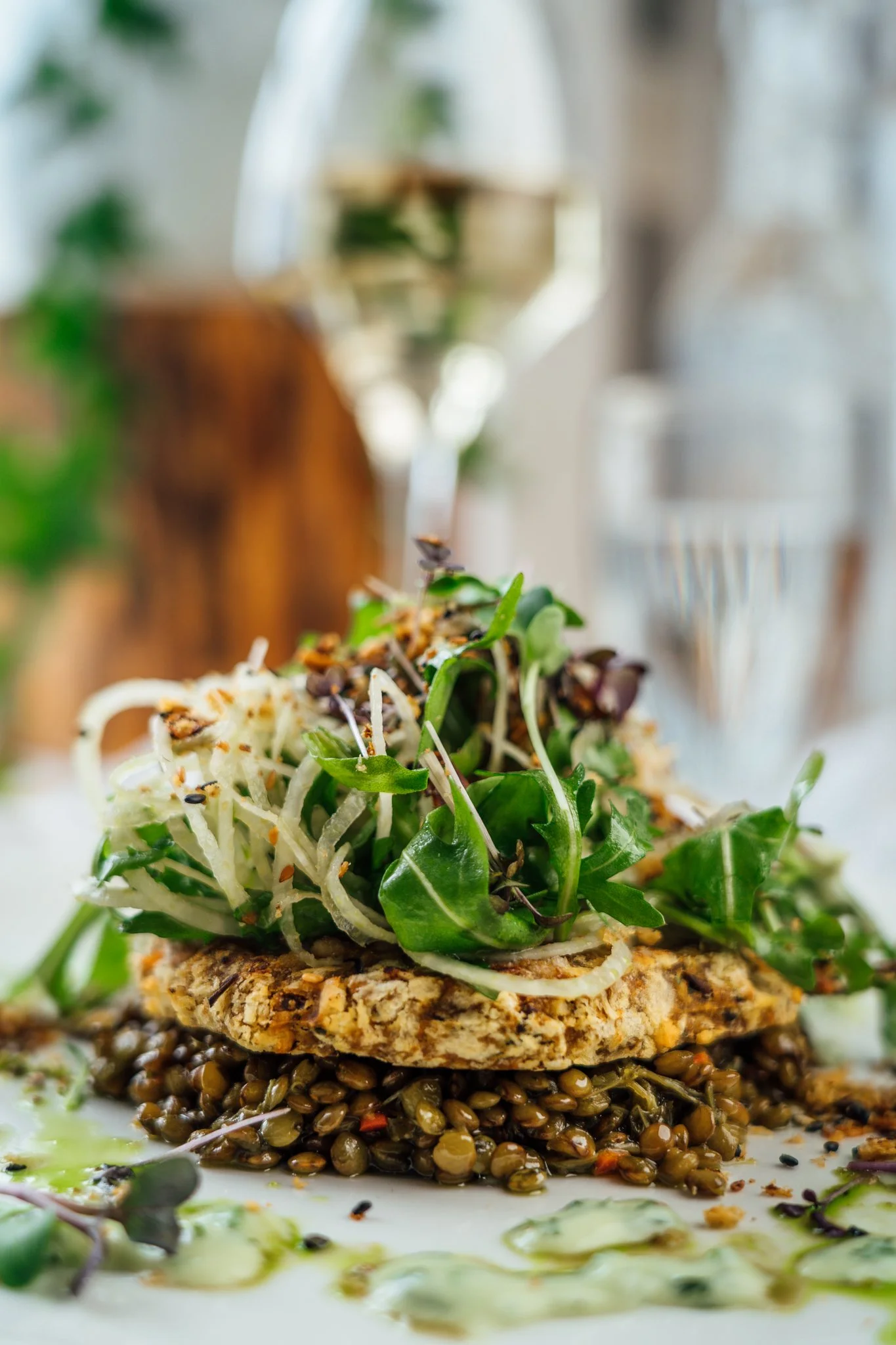 Close-up of a gourmet dish with a toasted grain patty, topped with greens and shredded cheese, served on a white plate with a glass of white wine in the background.