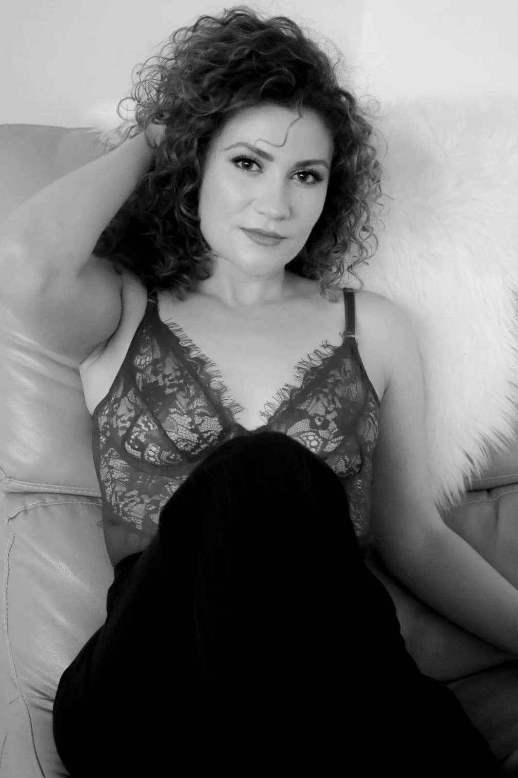 Lauren Keating wearing a lace top, sitting on a sofa with a furry pillow behind her, and a person with dark hair in the foreground.