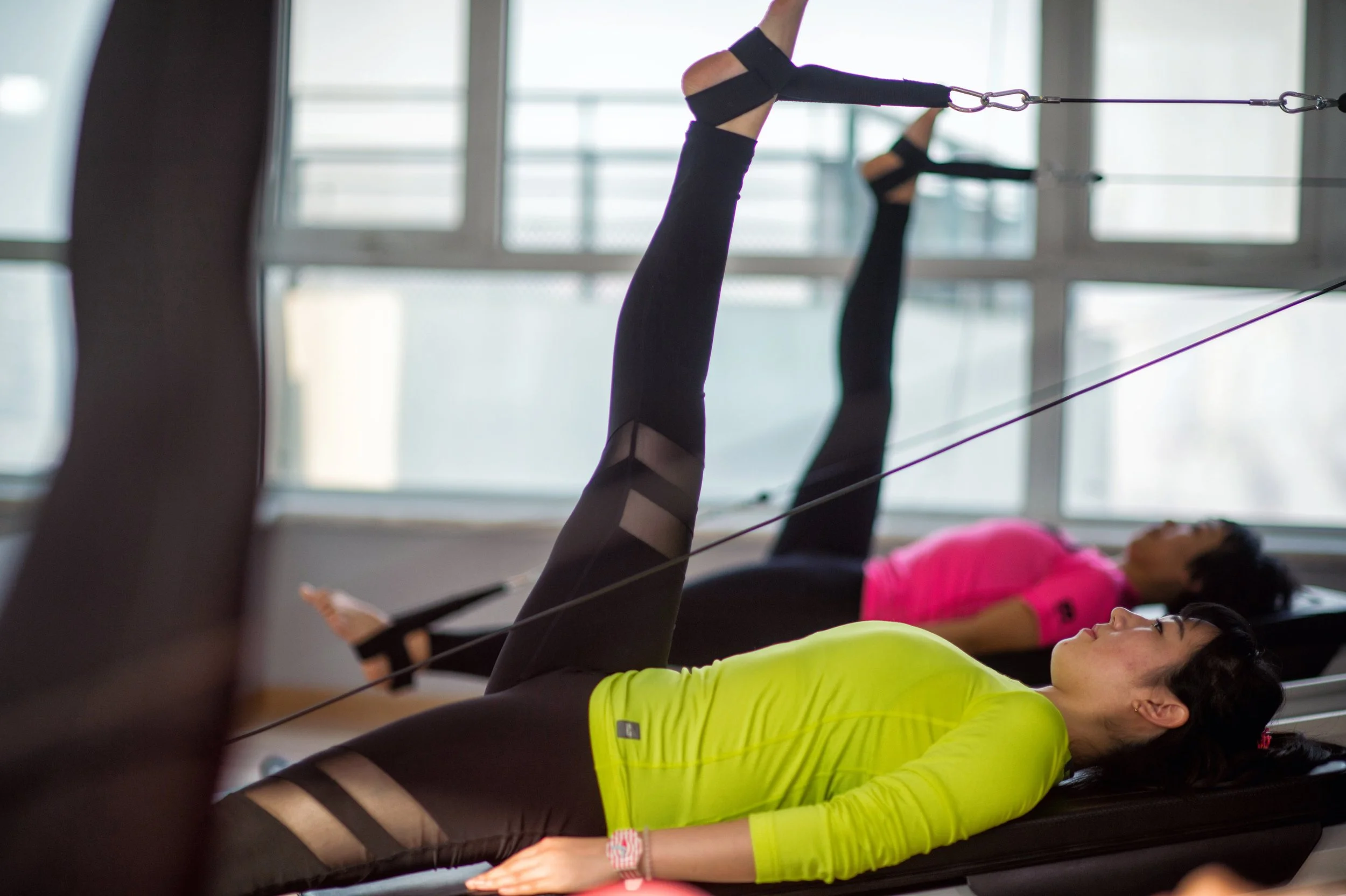 Two women lying on exercise mats in a gym, stretching their legs upwards while holding onto resistance bands attached to the ceiling.