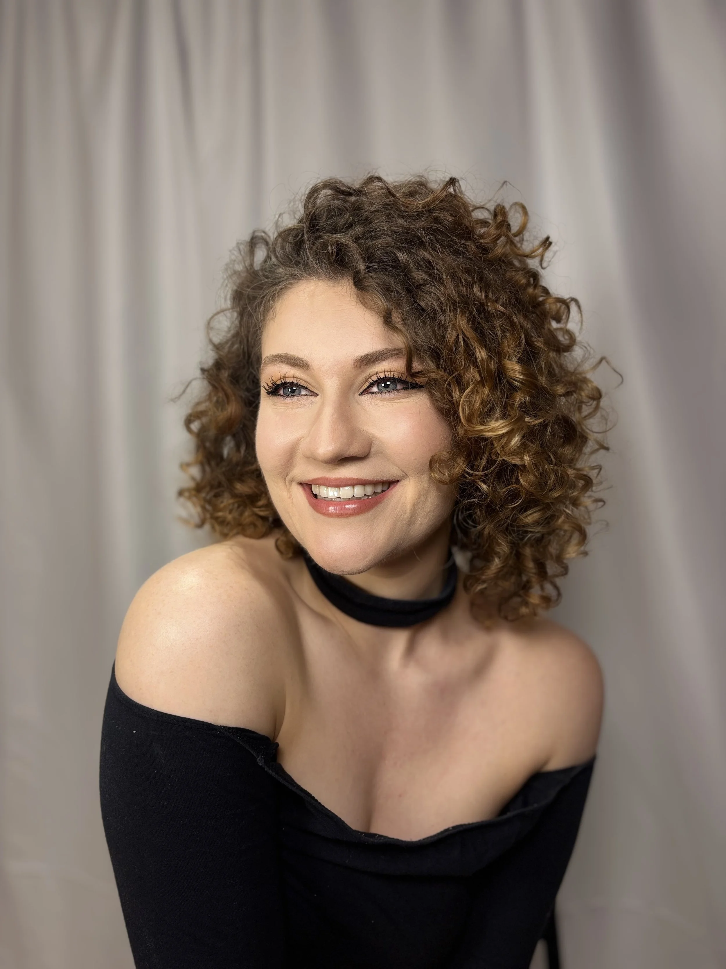 Lauren Keating smiling, wearing a black off-shoulder top and a black choker, against a gray curtain background.
