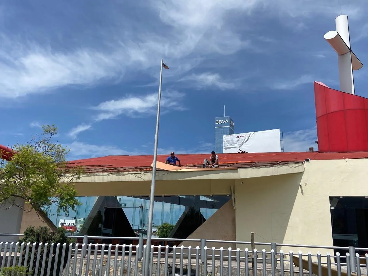 Dos trabajadores en el techo de un edificio con estructura moderna y ventanas de vidrio, debajo una cerca metálica y un árbol, en el fondo un cielo azul con nubes y un edificio alto con el cartel BBVA.