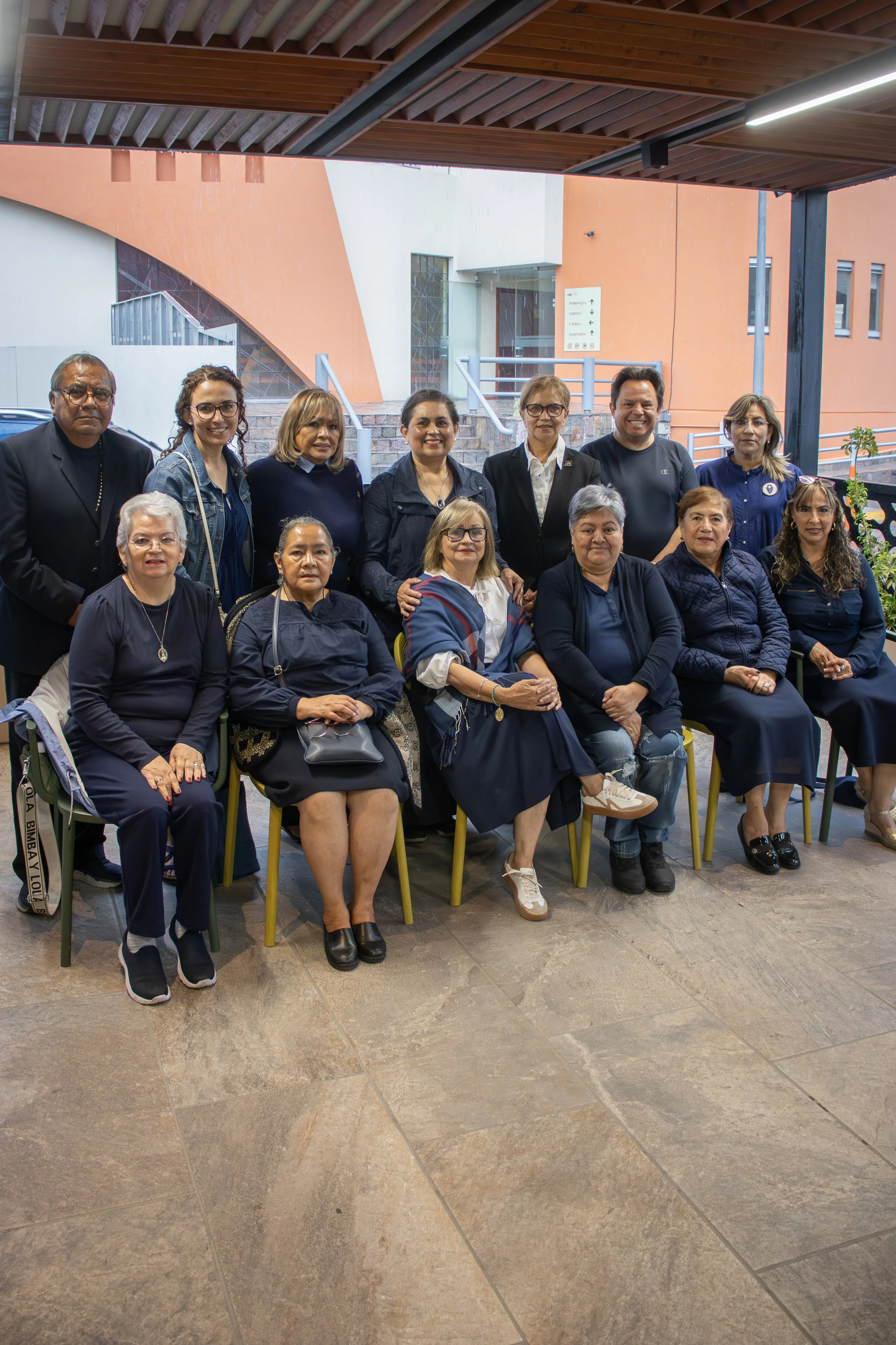 Grupo de personas posando para una foto en un lugar cubierto con estructura de madera, algunas sentadas y otras de pie, todas vestidas en tonos oscuros, en un ambiente exterior con un edificio en el fondo.