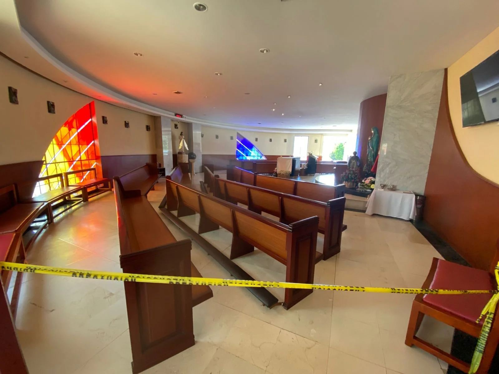 Sala de espera en un lugar religioso con asientos de madera, una estatua y una cruz en el fondo, y cinta de advertencia en primer plano.