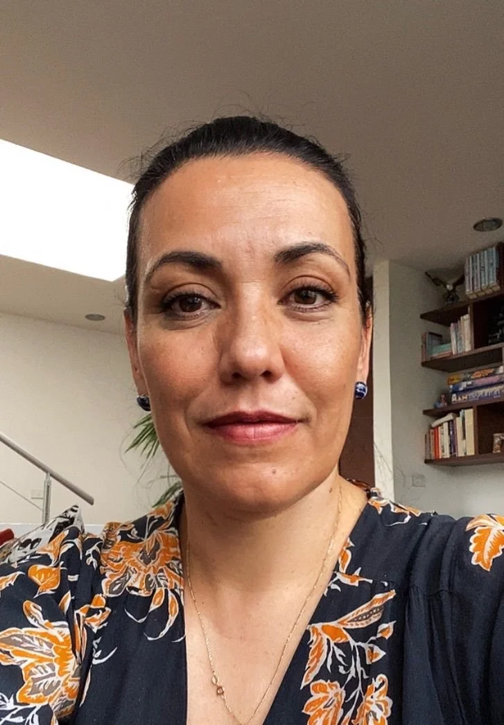 Retrato de una mujer con cabello recogido, usando un vestido oscuro con estampado de flores en tonos naranjas y blancos, en un interior con estanterías con libros.