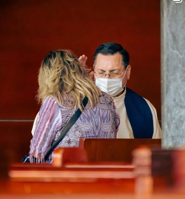 Una mujer y un hombre en una iglesia, la mujer le está susurrando, él lleva cubrebocas y parece molesto.