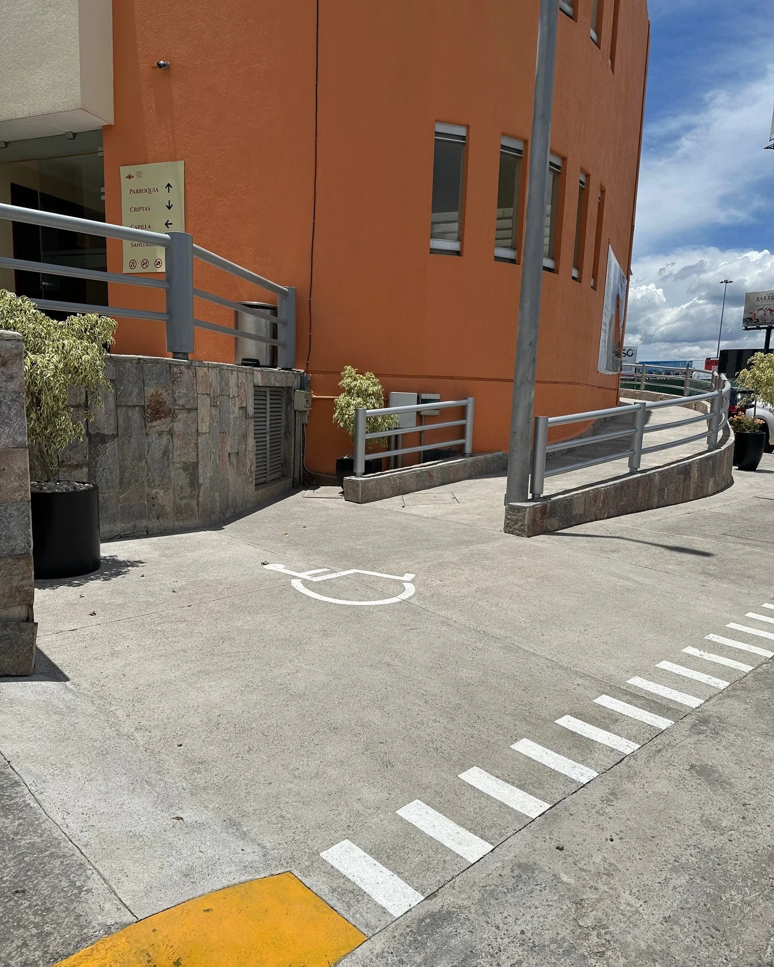 Estacionamiento con espacio para discapacitados con señal de acceso en el suelo y barandillas metálicas alrededor, cerca de un edificio de color naranja con ventanas.