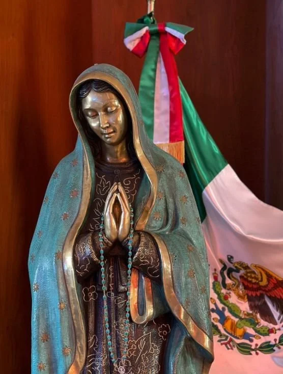 Estatua de la Virgen María con las manos juntas en oración, vestida con un manto azul y detalles dorados, frente a una bandera de México