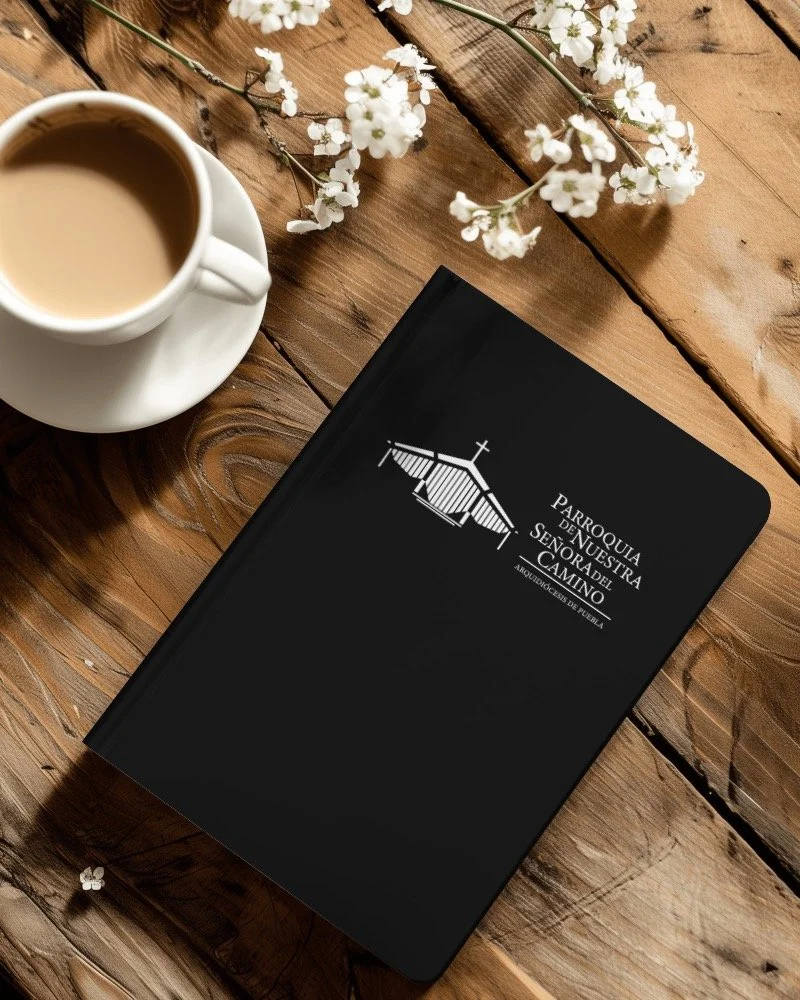 Una taza de café con leche, un libro negro con texto en blanco y una ilustración de una iglesia, y flores blancas sobre una mesa de madera.