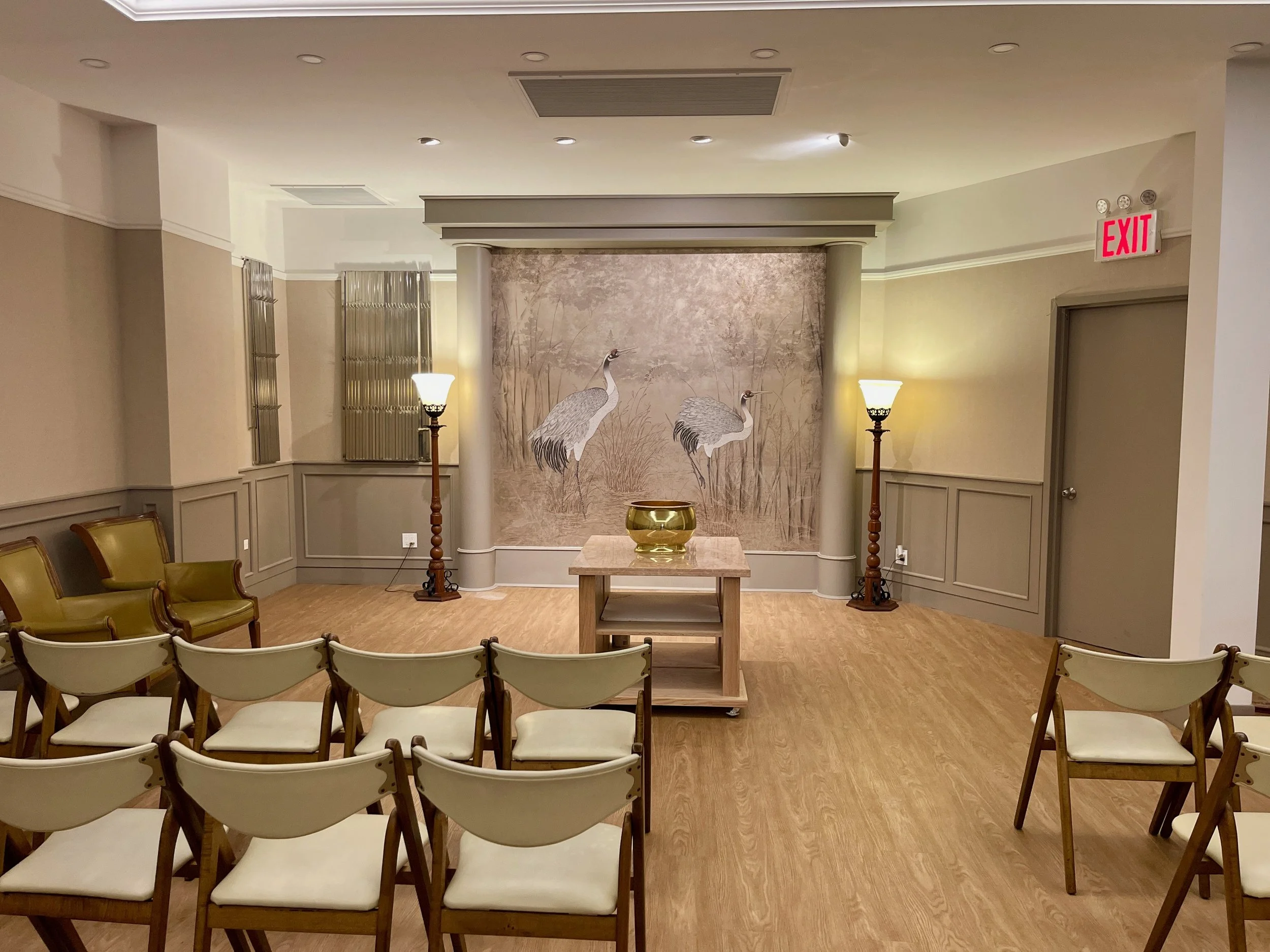 A funeral chapel with a backdrop of two cranes on a beige wall, surrounded by four beige chairs with wooden frames, two brown armchairs, two tall floor lamps, and a small table with a decorative bowl.