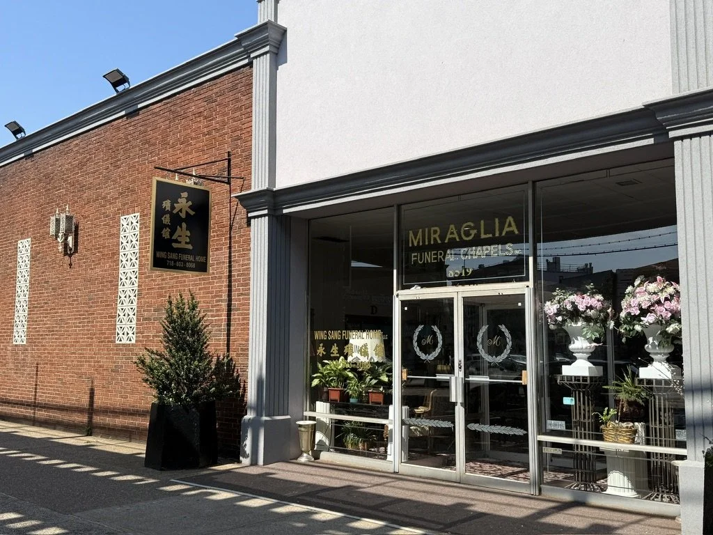 The entrance of a funeral home named Wing Sang Funeral Home, with the name displayed on the glass door. The building has a brick and white exterior with decorative potted plants and flowers outside.