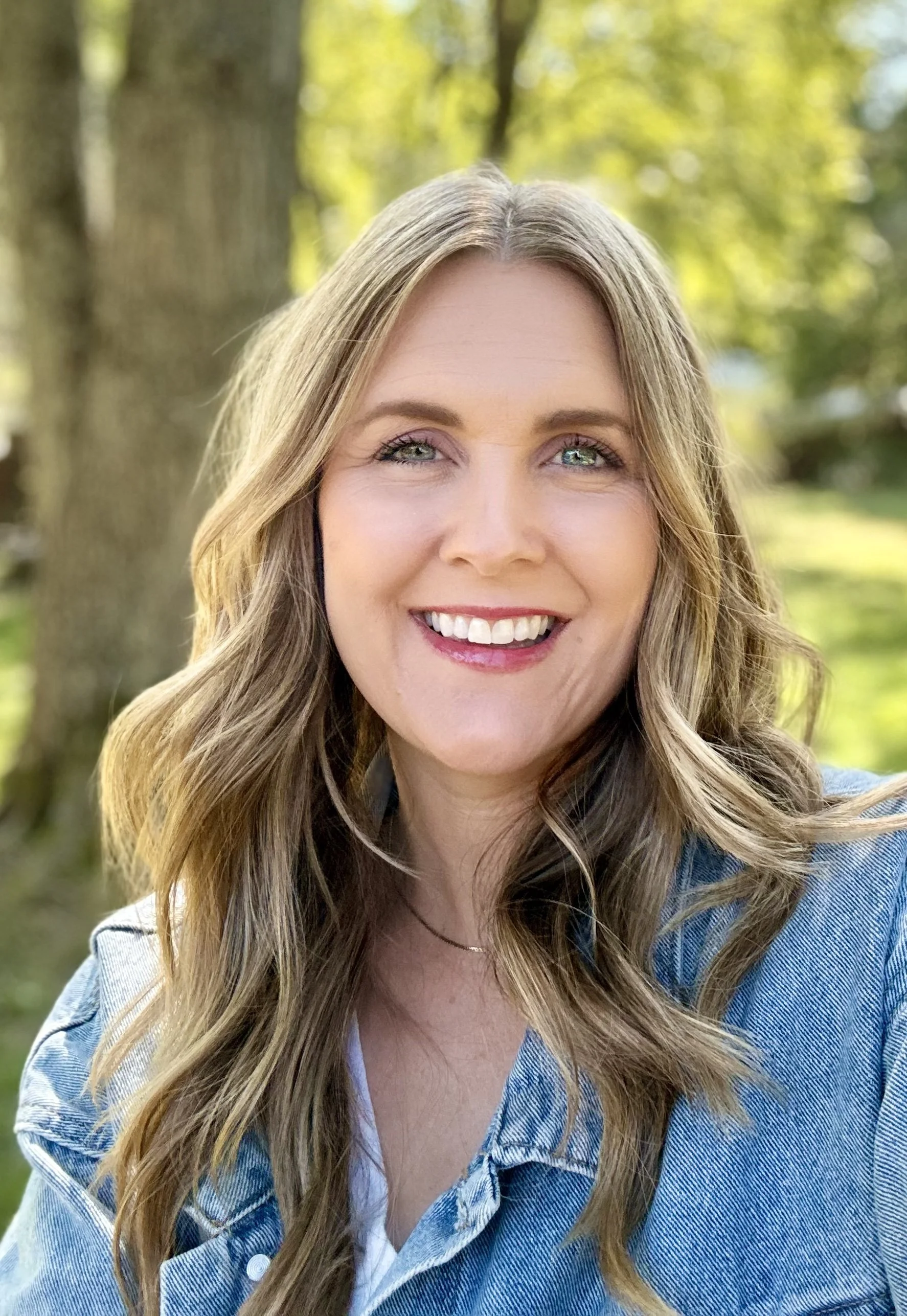 A woman with long wavy light brown hair and blue eyes smiling outdoors in a park or forest with green trees and sunlight in the background.