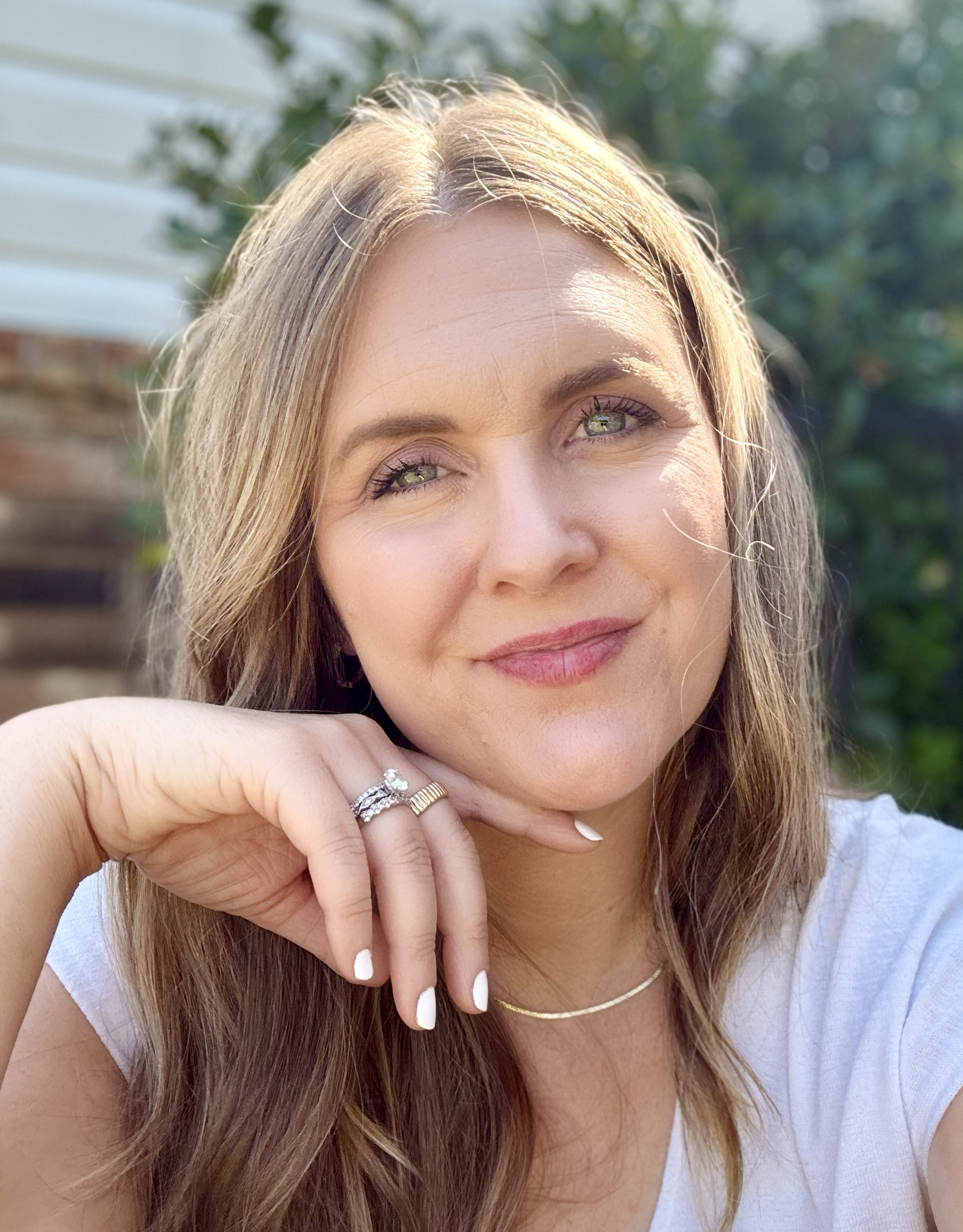 A woman with light brown hair and green eyes resting her chin on her hand, wearing rings, a necklace, and a white top, outdoors with greenery and a wooden fence in the background.