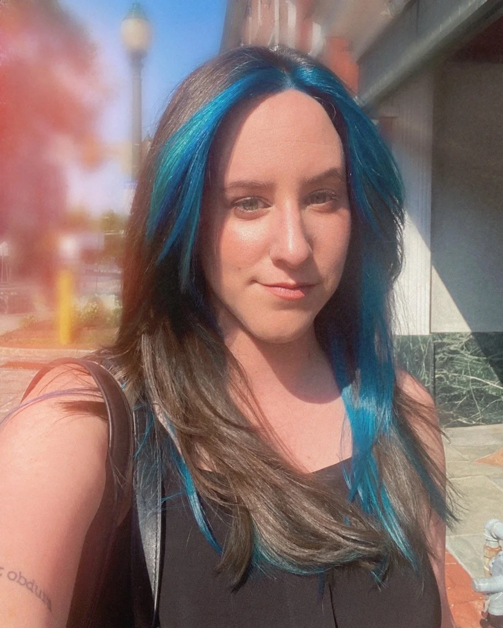 A woman with long dark brown hair streaked with bright blue highlights takes a selfie outdoors in sunlight, wearing a black top with a bag strap over her shoulder.