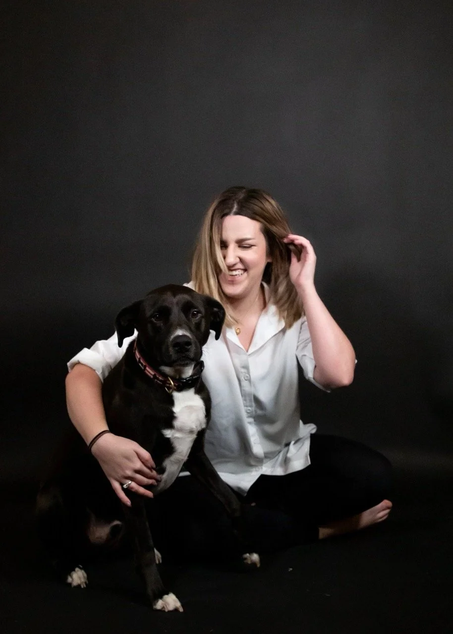A woman in a white shirt sits against a dark background, smiling with her hand in her hair, next to a large black dog with a white chest.