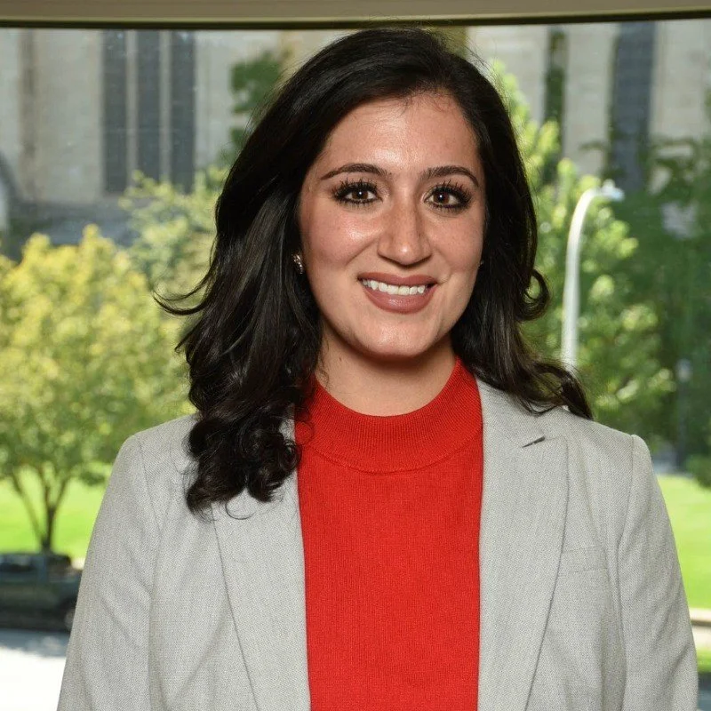A woman with shoulder-length dark hair wearing a red top and a light-colored blazer, standing indoors with a large window behind her showing trees and a building outside.