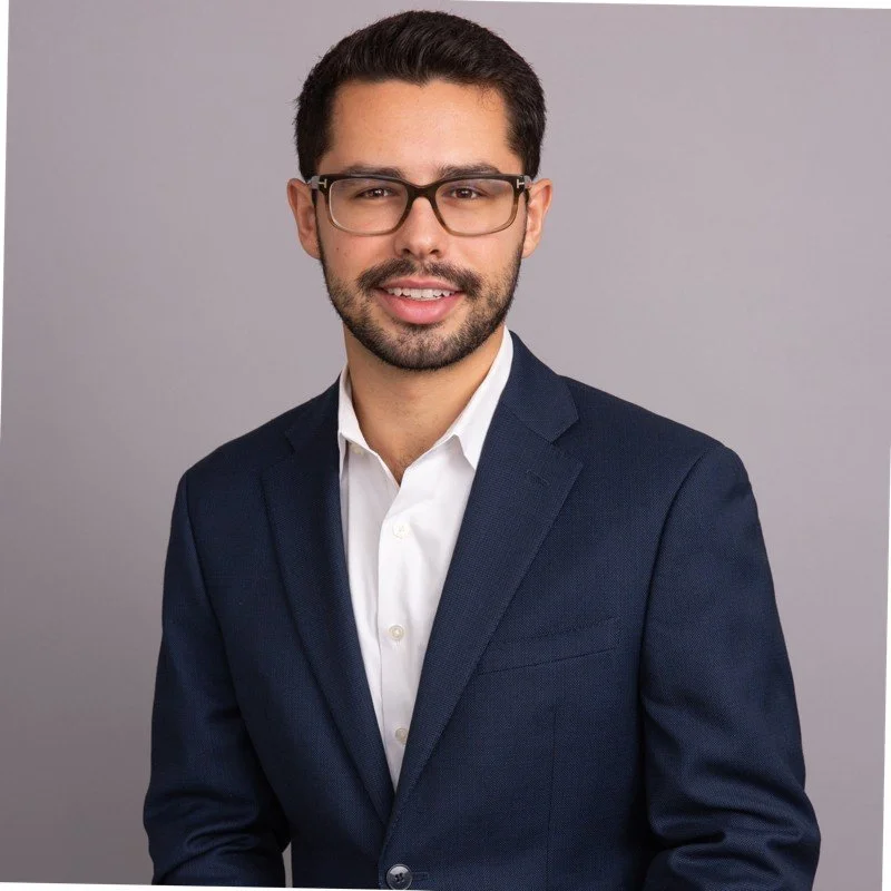 A young man with dark hair, glasses, a neatly trimmed beard, and a friendly smile, wearing a dark blue suit and white shirt, standing in front of a plain gray background.