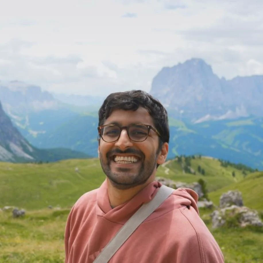Smiling man with glasses in a pink hoodie outdoors with green hills and mountains in the background.