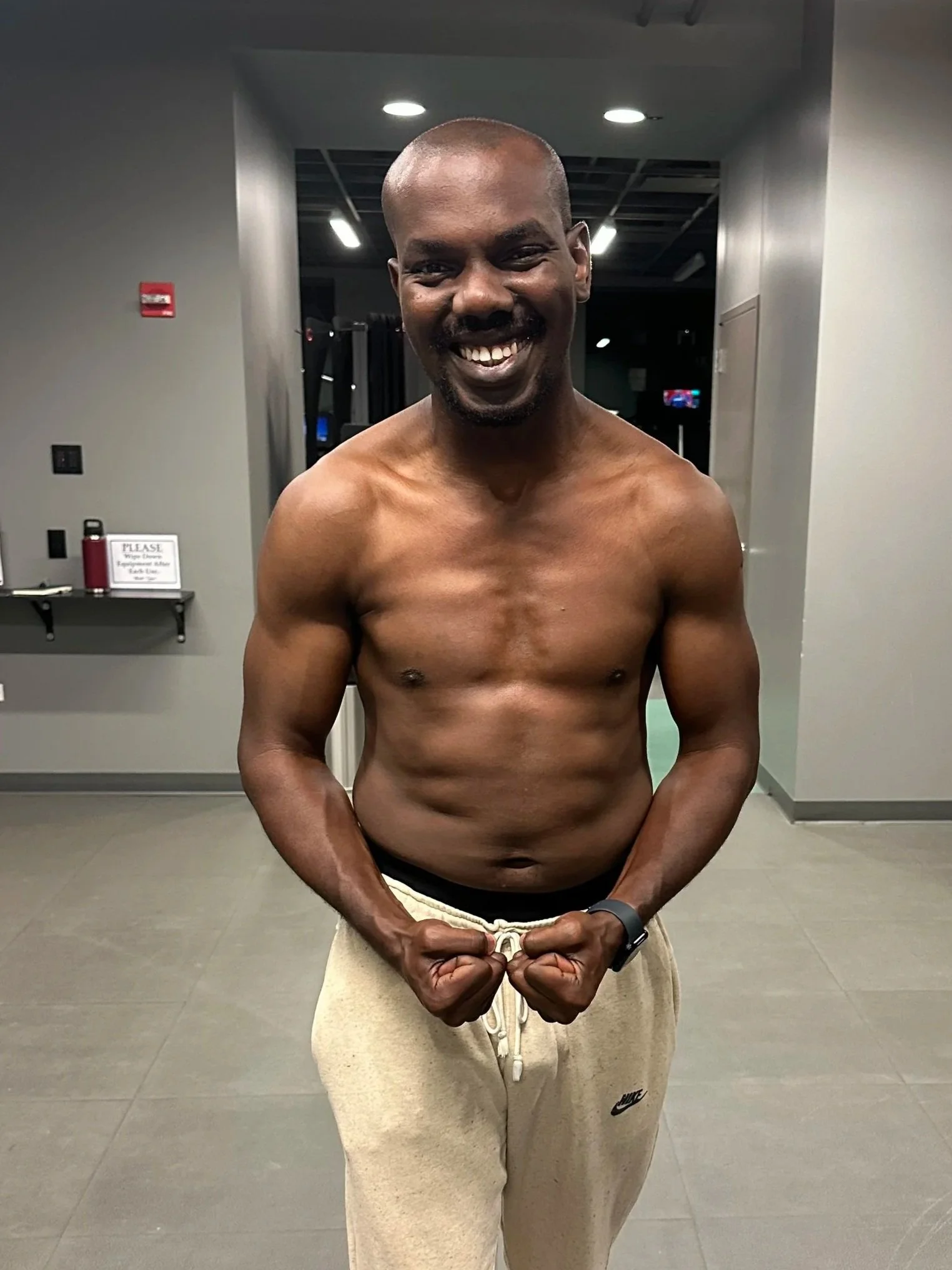 A shirtless man with a broad smile, standing in a gym, flexing his muscles.