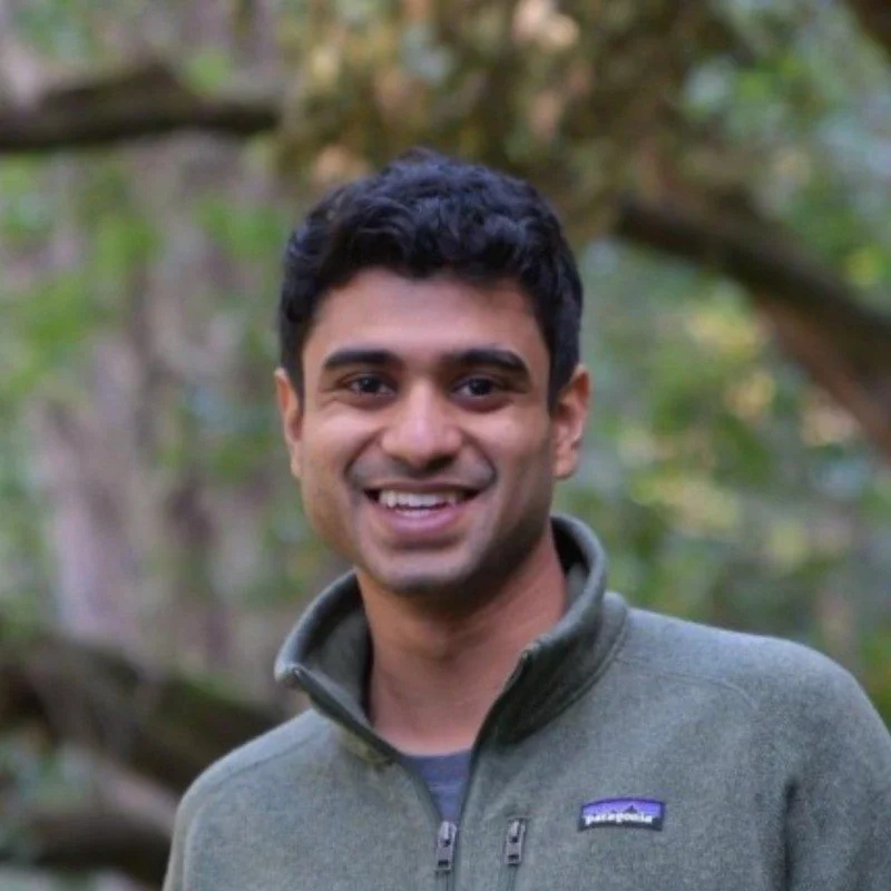 A smiling man with dark hair, wearing a green fleece jacket, standing outdoors with trees and blurred greenery in the background.