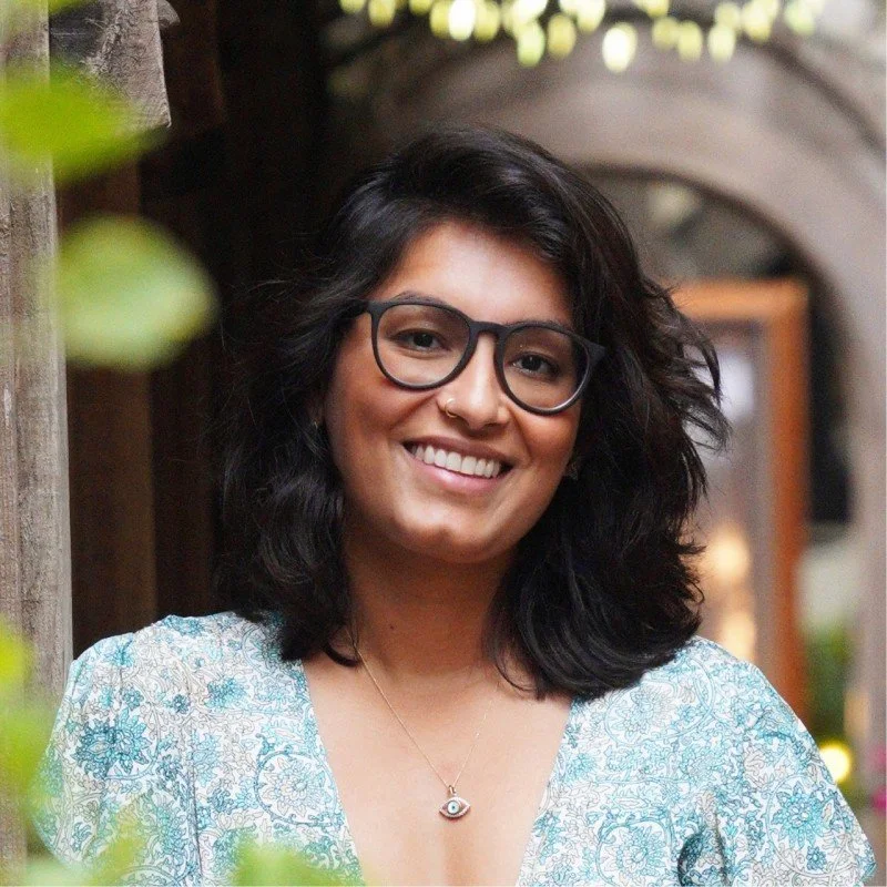 A young woman with shoulder-length black hair, glasses, and a nose ring, smiling outdoors near a wooden structure with string lights overhead.
