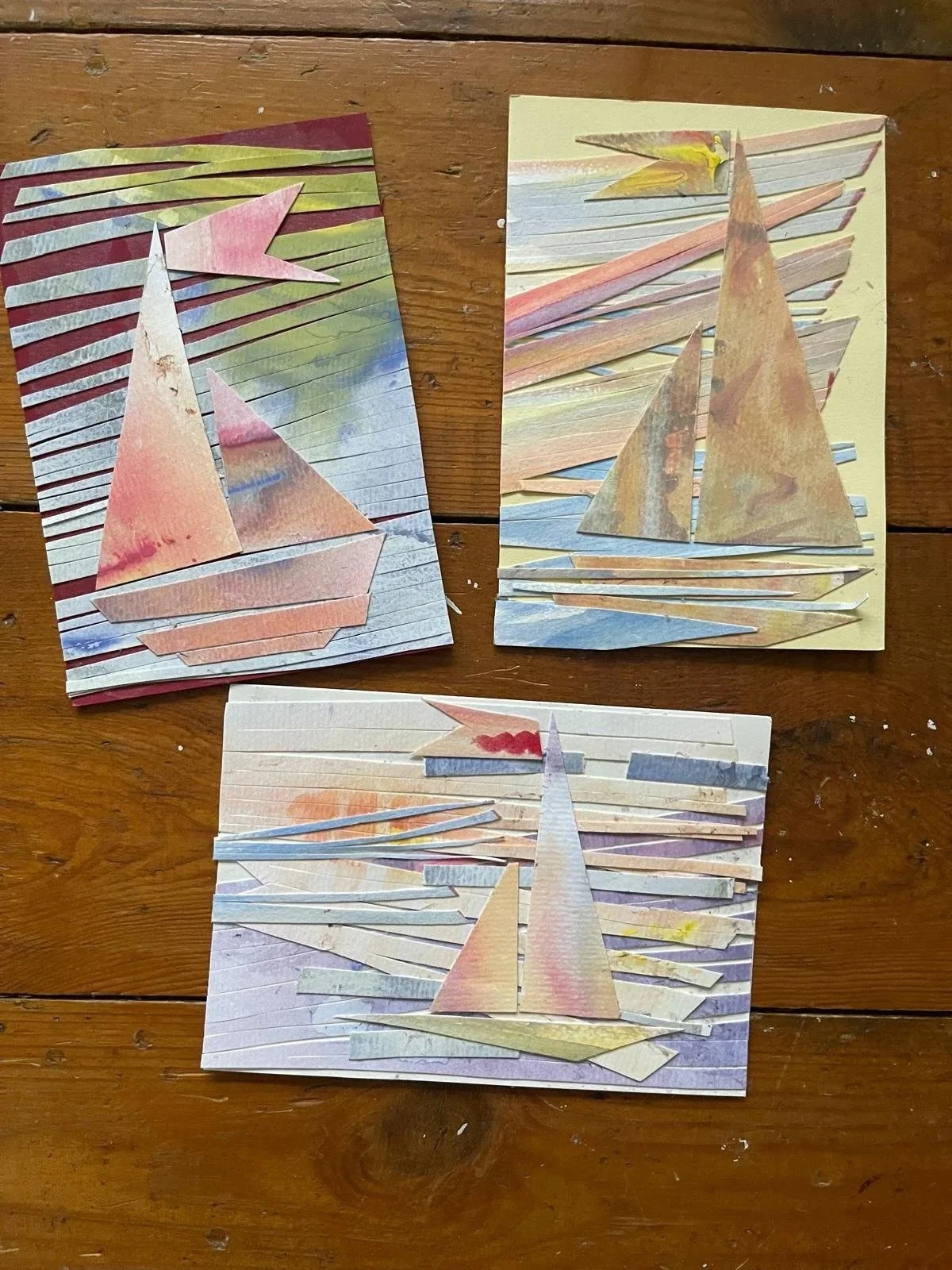 Three pieces of artwork made from cut and layered pieces of colored paper arranged on a wooden surface, creating abstract, geometric designs.