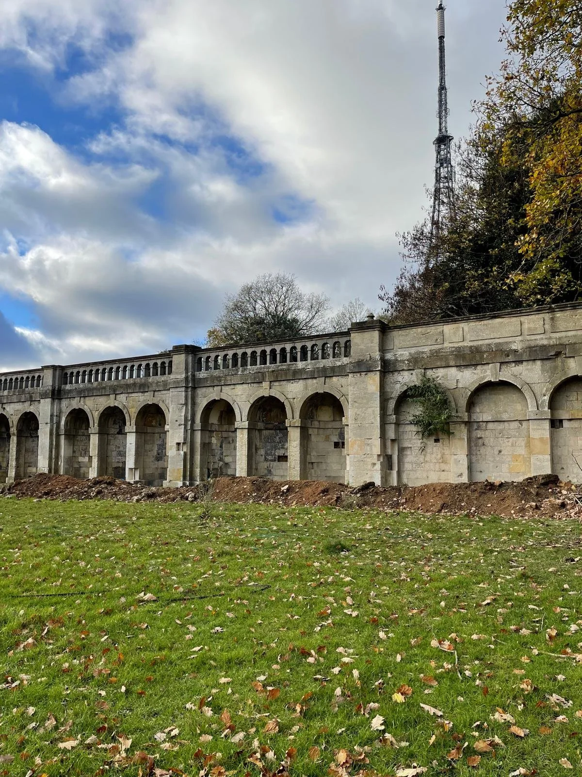 Old stone wall with arches, construction debris at the base, green grassy area with fallen leaves, trees on either side, blue sky with clouds, communication tower above the wall.