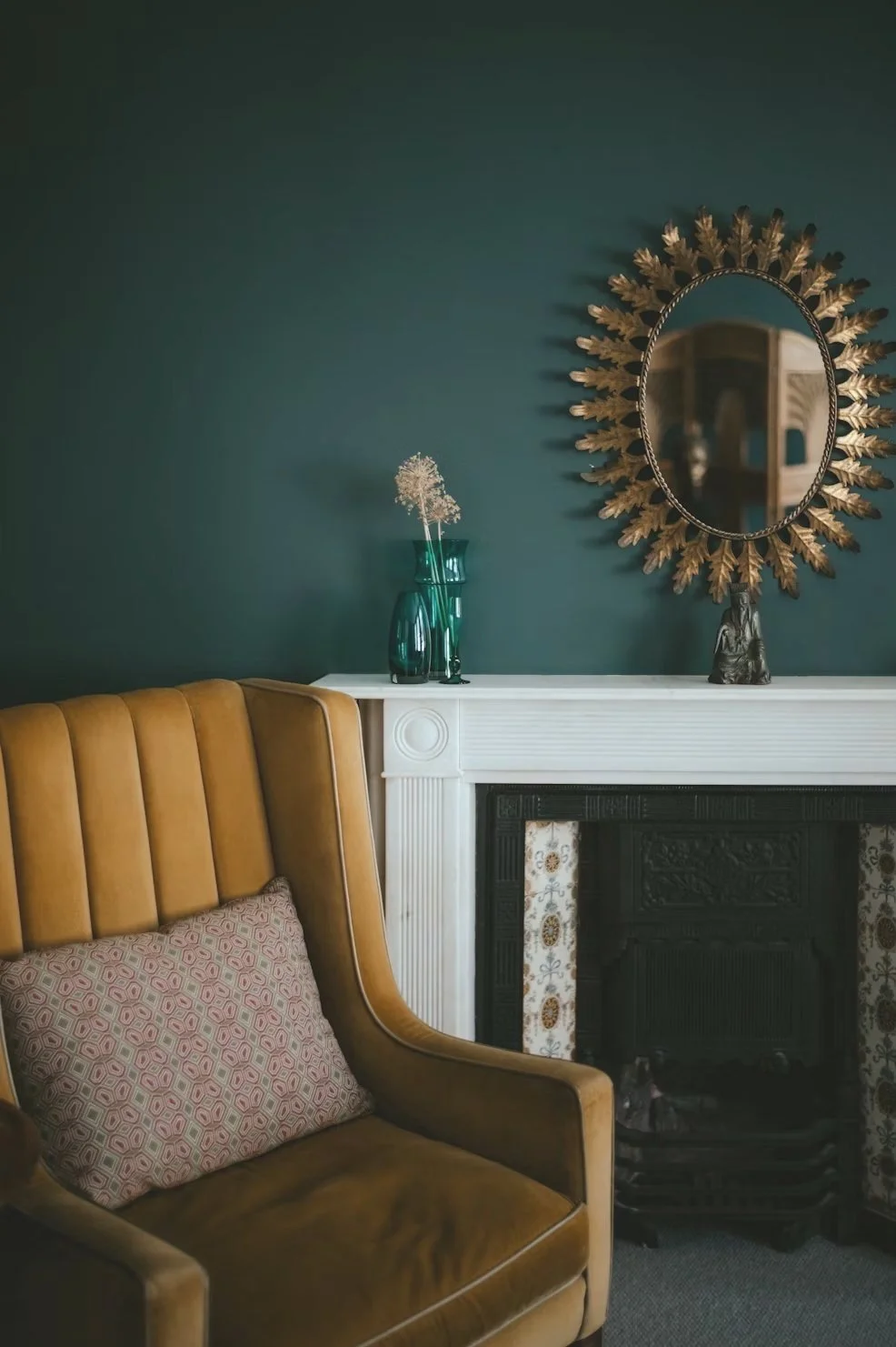 Living room with a mustard yellow armchair, a white fireplace mantel, a decorative mirror with a sunburst frame, a small brown sculpture, and two dark green vases with dried flowers on the mantel. The wall is painted teal.