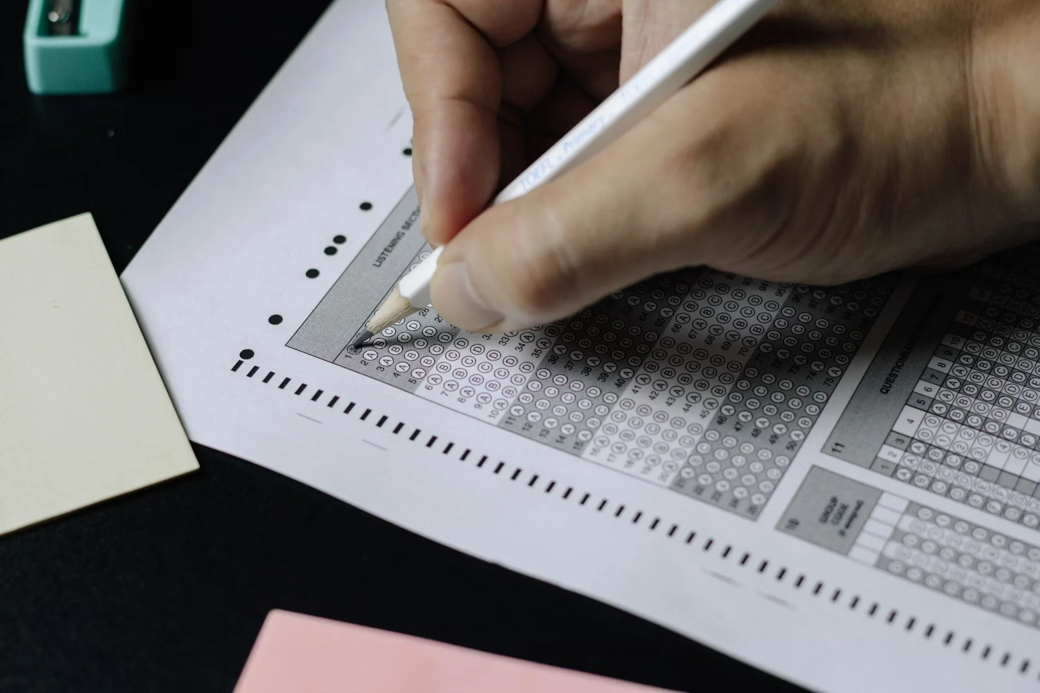 Close-up of a hand filling out an answer sheet with a pencil, with colored sticky notes and study supplies nearby.