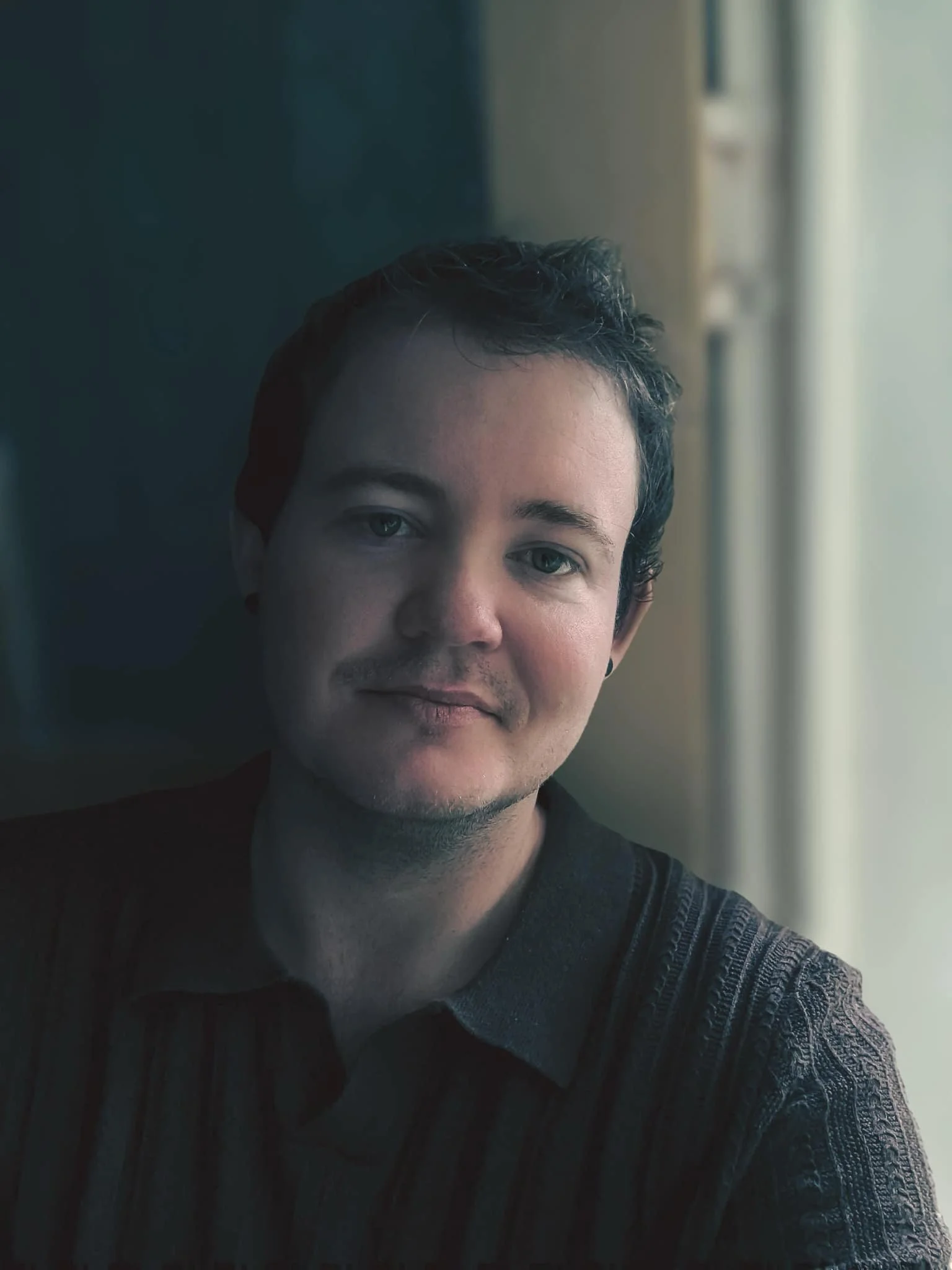 A young man with short dark hair and light skin sitting near a window, looking at the camera with a slight smile, wearing a dark patterned shirt, with natural light illuminating his face.