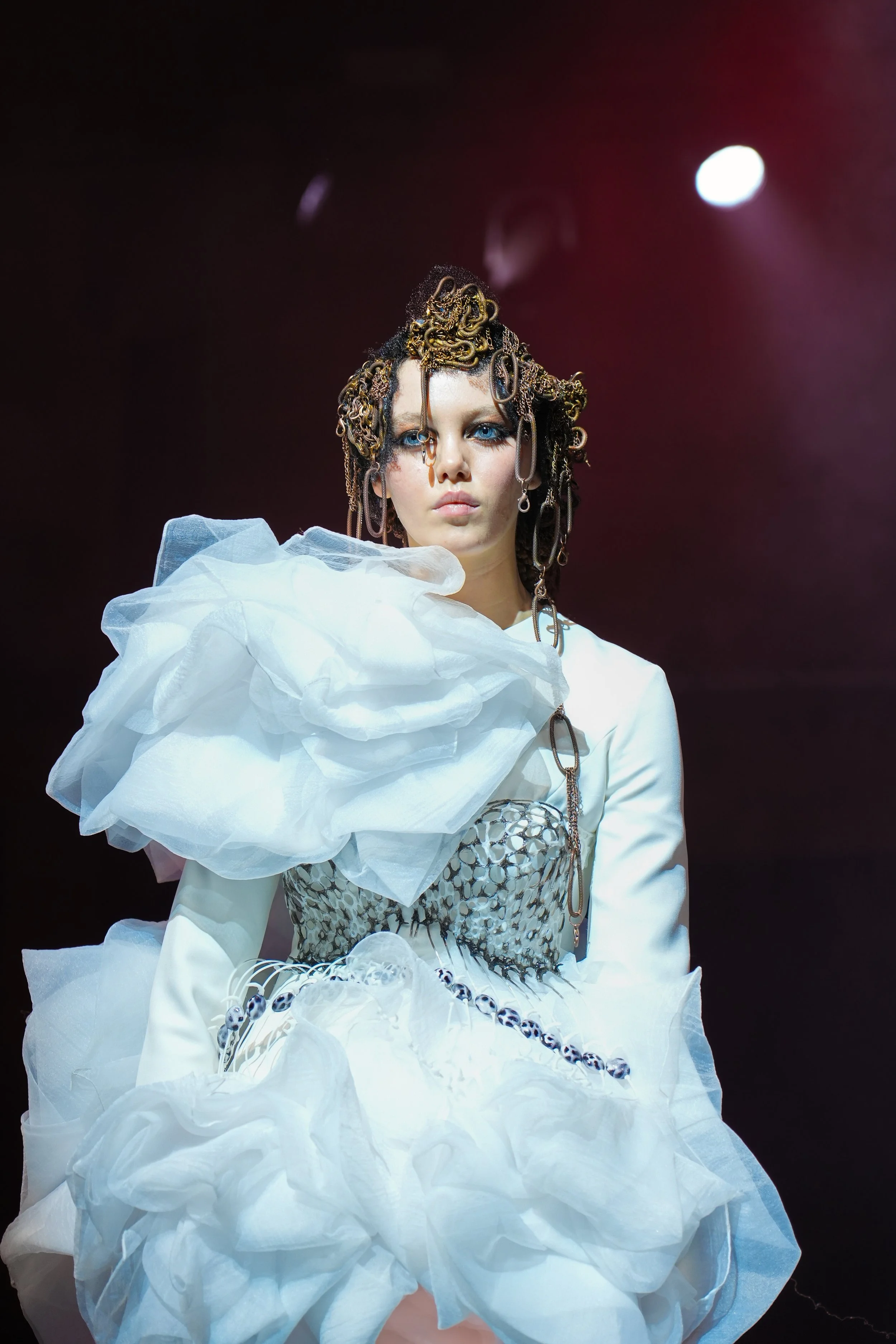 Model walking on runway wearing a white avant-garde dress with ruffled details and metallic accents, with hair styled in buildup of curly braids and chains, under stage lighting.