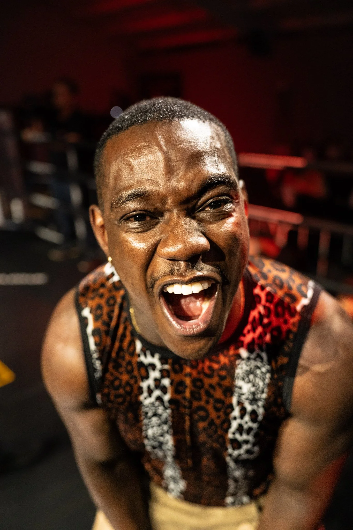 A young man with a short haircut and dark skin, smiling widely with mouth open, wearing a sleeveless animal print shirt, in an indoor setting with a blurred background.