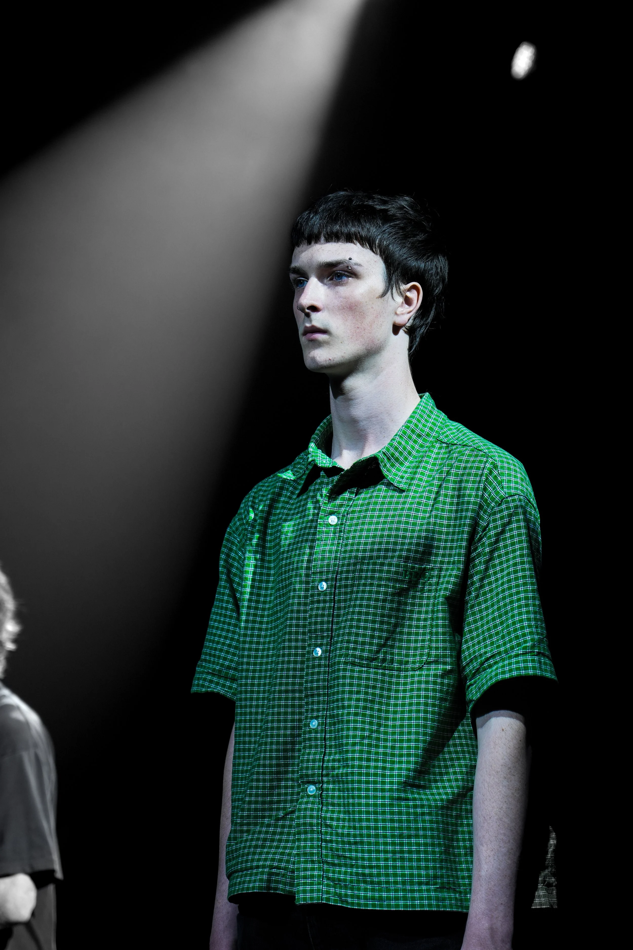 A young man with short dark hair, wearing a green checkered shirt, standing on a dark stage with a spotlight shining down on him.