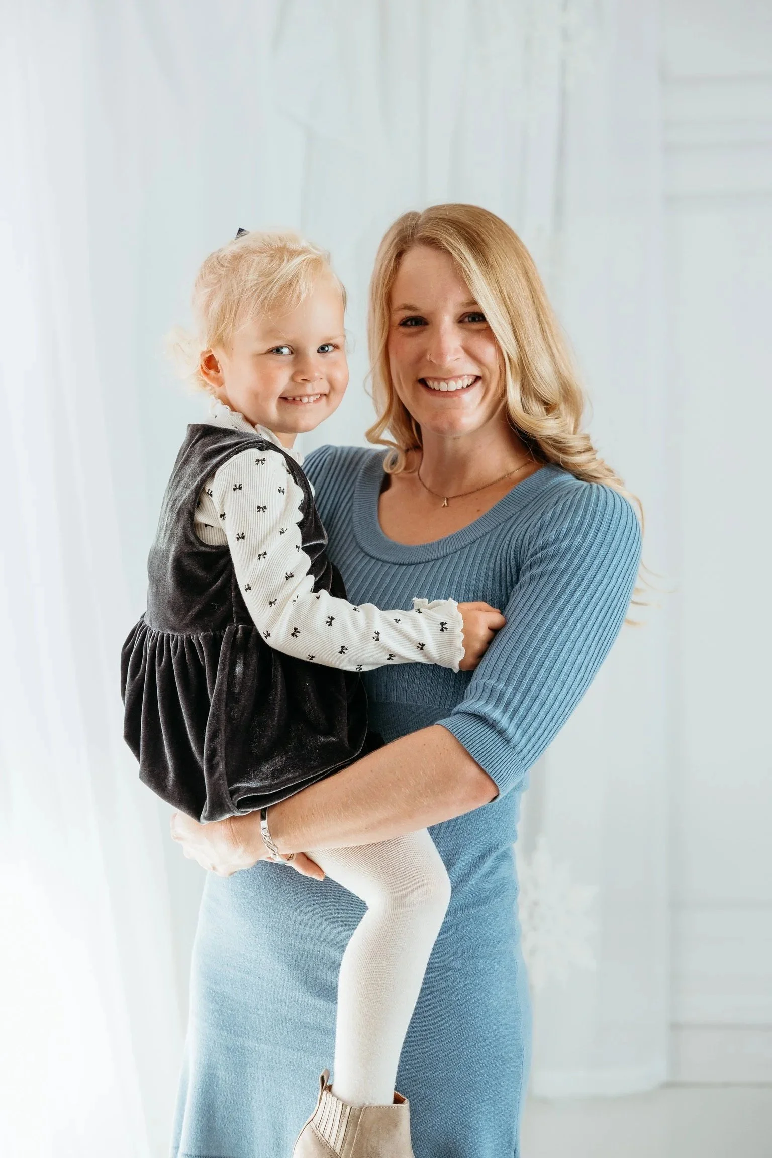 A woman with blonde hair holding a young girl with blonde hair, both smiling at the camera indoors.