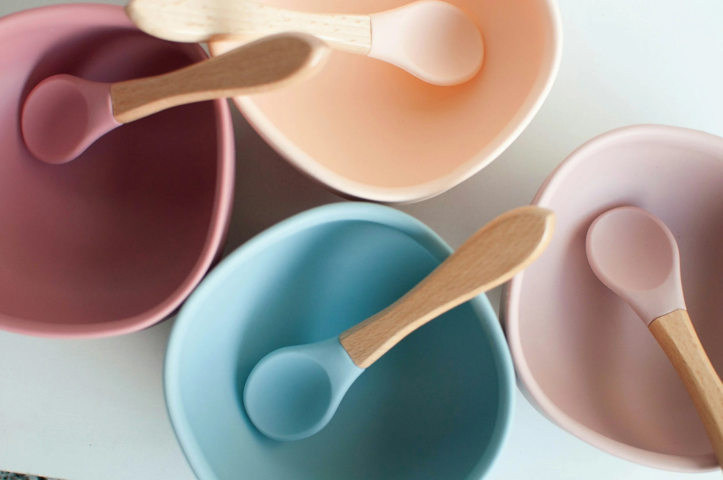 Colorful ceramic bowls and silicone spoons with wooden handles arranged on a white surface.